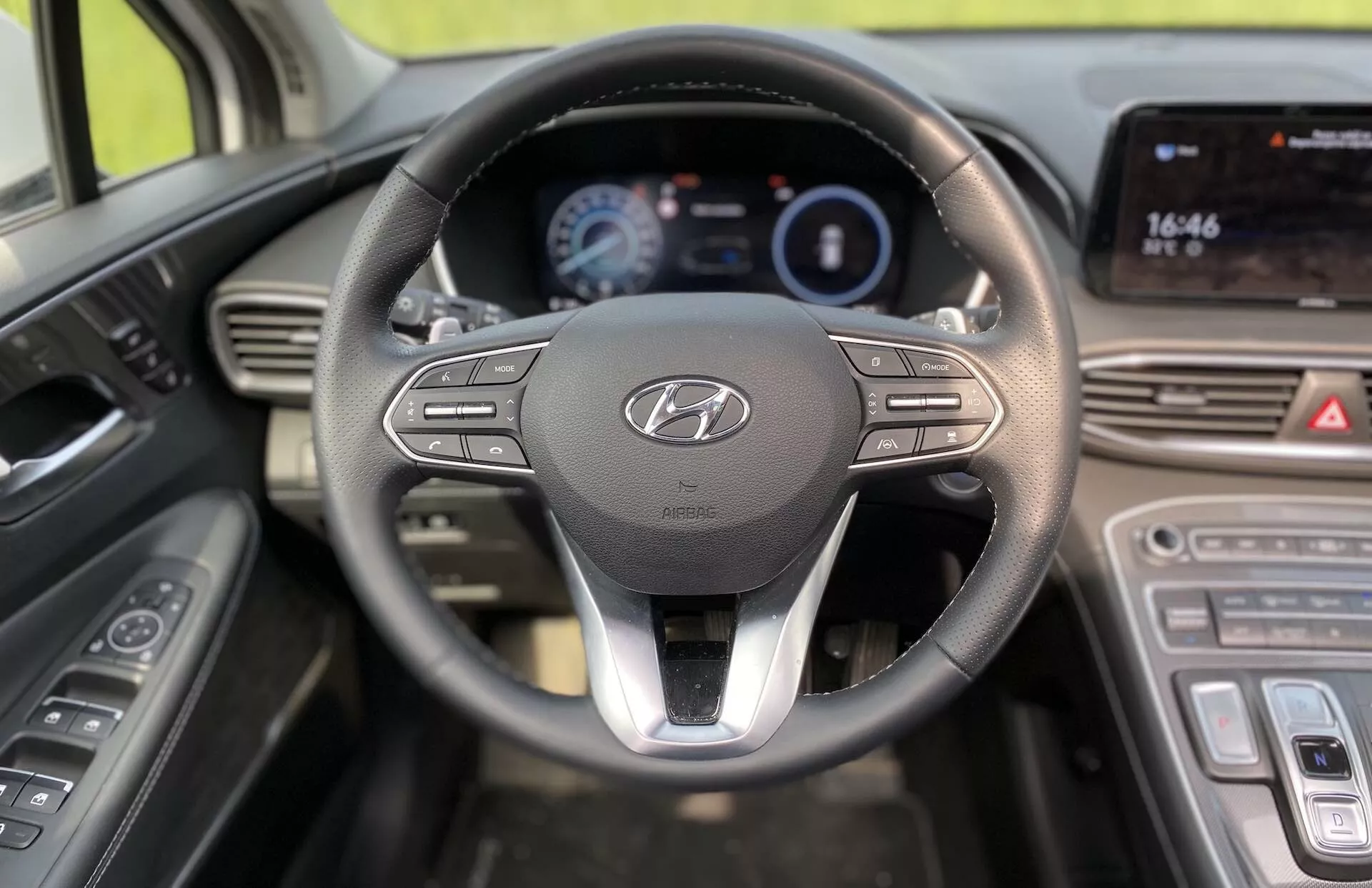 Perspective du conducteur de l'intérieur d'une Hyundai Santa-Fe 2020 modèle hybride, avec un volant proéminent arborant le logo Hyundai et des boutons de commande pour le multimédia et la connectivité. Derrière le volant se trouve un tableau de bord numérique éclairé affichant des informations clés telles que la vitesse et la consommation de carburant. À droite, la console centrale comprend un écran tactile affichant l'heure (16:46) et probablement des fonctionnalités de navigation et multimédia. Sous l'écran tactile, des commandes de climatisation tactiles sont visibles, renforçant le design moderne du véhicule. Les commandes des portes pour les vitres électriques et les réglages des rétroviseurs latéraux sont visibles à gauche du volant.