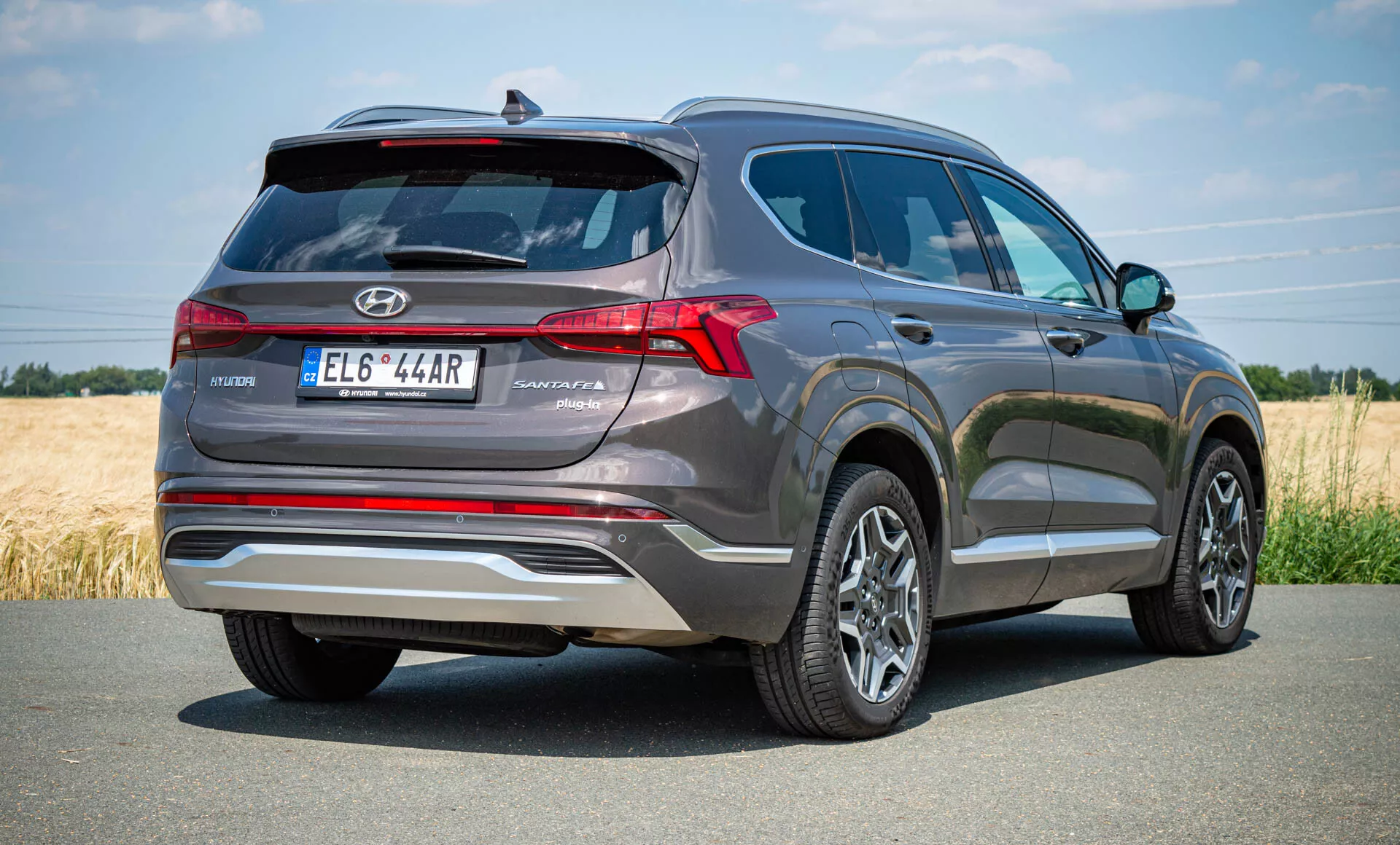 Vue arrière trois quarts d'une Hyundai Santa Fe Hybrid 2020. La prise de vue en contre-plongée met en valeur la hauteur et la robustesse du SUV, avec la voiture positionnée sur le côté droit suggérant un mouvement. Garée sur une route pavée dans un cadre rural, l'arrière présente des feux arrière à LED disposés verticalement, un spoiler arrière pour la stabilité aérodynamique, des doubles sorties d'échappement, un spoiler de toit fractionné et des garnitures réfléchissantes sur le pare-chocs. Le design laisse entrevoir des capteurs de stationnement arrière, une caméra de recul, des roues en alliage multi-branches, et une antenne aileron de requin pour la radio satellite ou la technologie GPS.