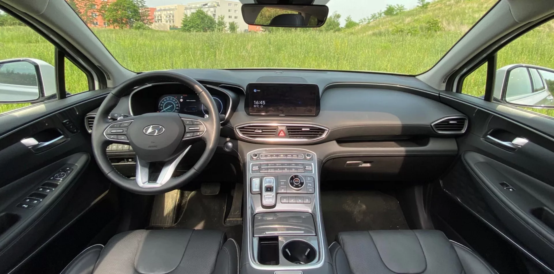 Vue intérieure du Hyundai Santa Fe 2020, montrant la cabine avant moderne avec un tableau de bord central doté d'un écran d'infodivertissement digital, une bande de ventilation élégante et des boutons multifonctions sur le volant. Le design du volant à gauche comprend le logo proéminent du constructeur et probablement un tableau de bord digital. La console centrale comporte un sélecteur de vitesse rotatif, des porte-gobelets et peut-être un accoudoir. Les sièges avant sont revêtus de cuir noir avec des coutures visibles, suggérant des réglages électriques et un luxe. Le cadre semble être extérieur avec une lumière naturelle, avec un champ vert légèrement flou visible à travers le pare-brise.