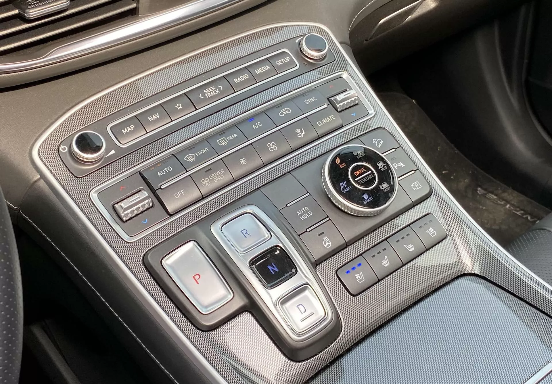 Cette image montre une vue rapprochée de la console centrale de la Hyundai Santa Fe 2020, se concentrant sur la commande de climat et la commande de transmission. La prise de vue, légèrement inclinée de la perspective du conducteur ou du passager, révèle le panneau de commande du climat disposé horizontalement en haut, comprenant les commandes de vitesse du ventilateur, les boutons de flux d'air, le contrôle automatique du climat, le dégivrage et la recirculation de l'air. En dessous, des boutons de sélection des vitesses avec un grand bouton 'P' pour le stationnement et un levier de type interrupteur pour les vitesses 'R' et 'N' sont présents, indiquant l'utilisation d'un système électronique ou à bouton pour la sélection des vitesses. Un cadran ronde pour la sélection des modes de conduite, entourés de boutons pour le frein auto-hold, l'assistant de stationnement et le système start/stop automatique est présent. Les commandes des sièges chauffants ou ventilés sont visibles en bas à droite. La console présente des matériaux premium avec des accents métalliques et un motif semblable à de la fibre de carbone. Un port USB en bas à gauche offre des options de connexion pour les dispositifs.