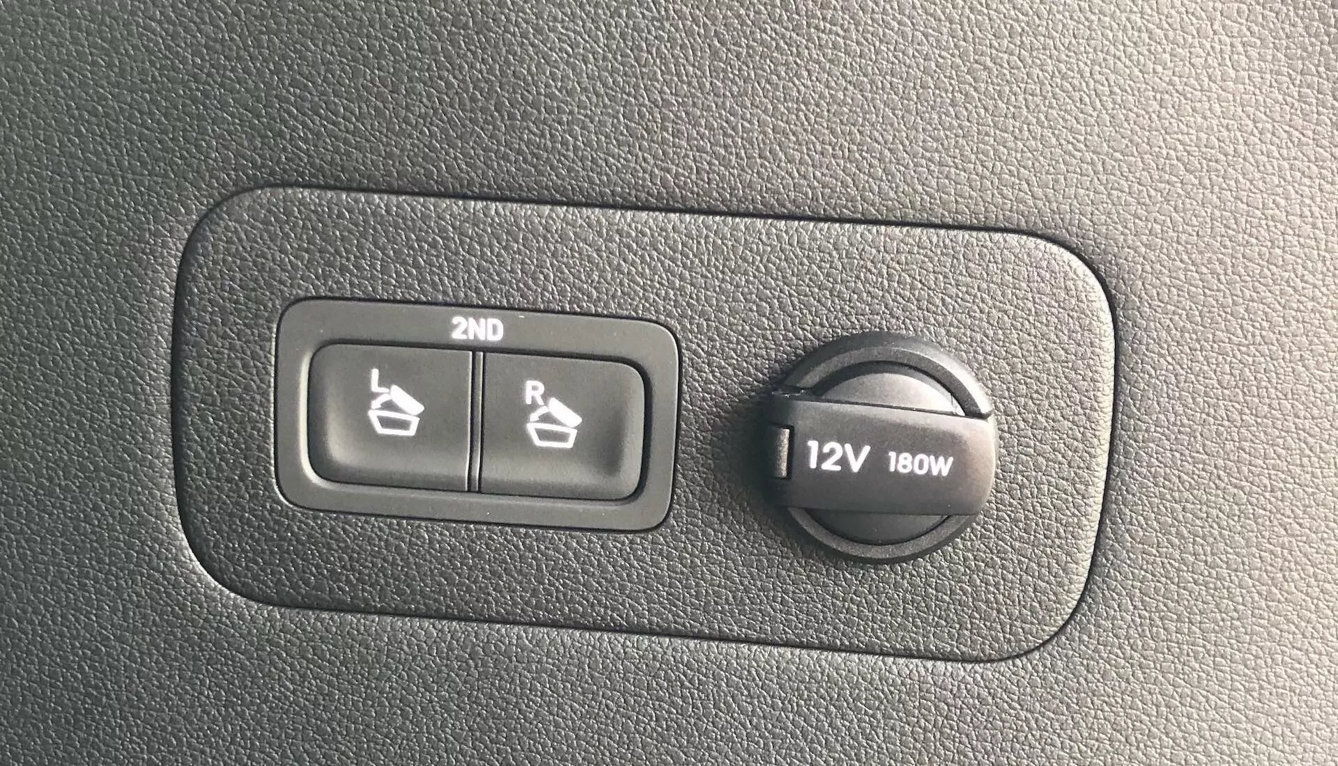 Gros plan sur une section du tableau de bord de la Hyundai Santa Fe 2020 montrant les commandes de chauffage des sièges pour la deuxième rangée et une prise de courant de 12V 180W sur une surface en plastique texturé.