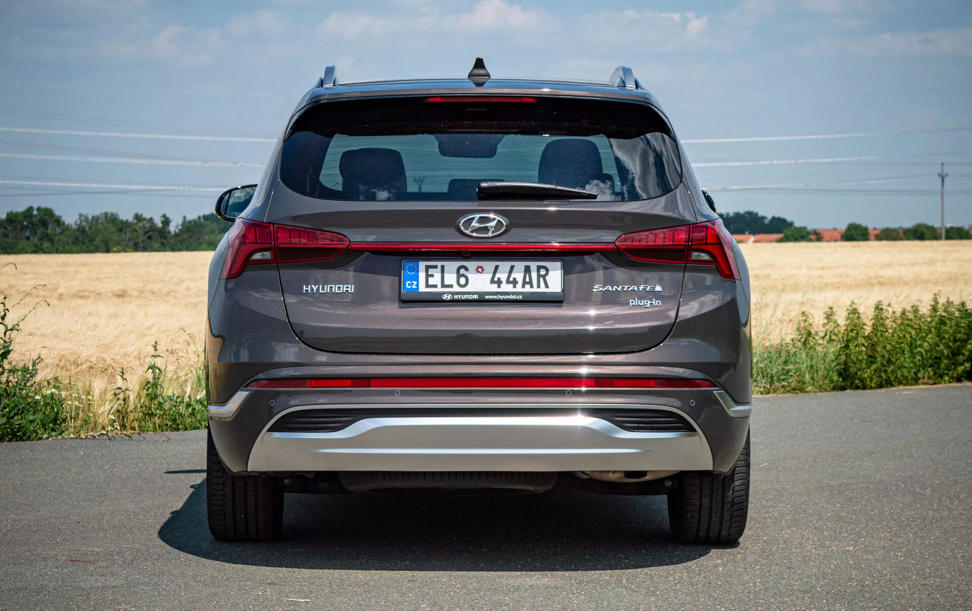 Vue arrière d'un SUV Hyundai Santa Fe 2020 gris argenté moderne stationné sur une route pavée, montrant le logo Hyundai, le nom du modèle Santa Fe, plaque d'immatriculation de style européen, feux arrière, pare-chocs arrière métallique réfléchissant avec capteurs de stationnement intégrés, tuyaux d'échappement doubles, vitre arrière avec becquet Hyundai et troisième feu stop. Temps clair avec un champ en arrière-plan.