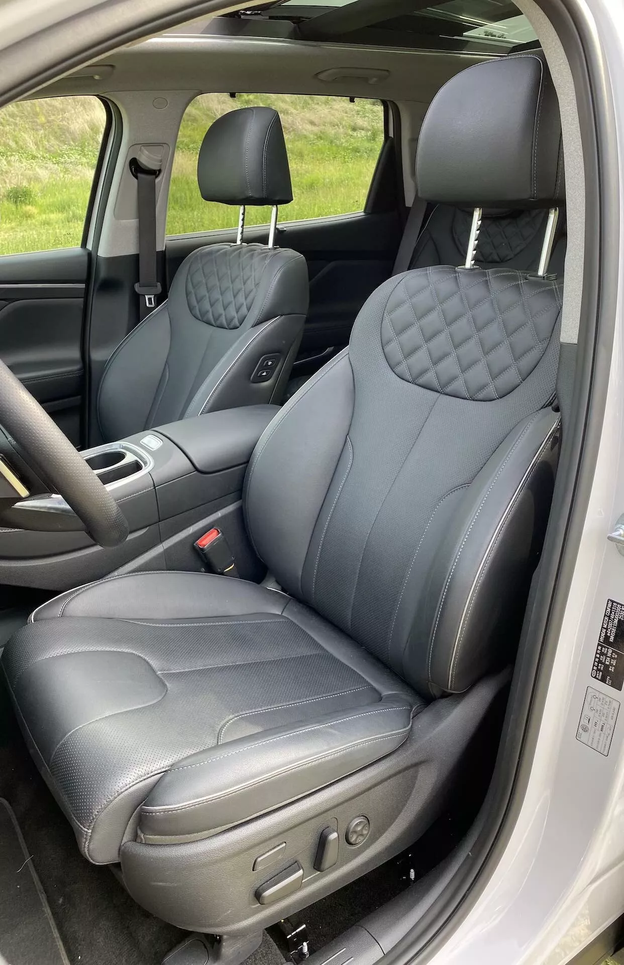 Intérieur du Hyundai Santa Fe 2020 montrant le siège du conducteur depuis une porte ouverte. La prise verticale en gros plan capture les sièges avant, en se concentrant sur le siège conducteur qui présente une sellerie en cuir foncé avec un motif de losanges matelassés sur le dossier et le coussin du siège, suggérant une variante premium. Les commandes électroniques visibles indiquent des positions de siège réglables. L’appuie-tête semble réglable et le panneau de porte comporte des commandes de vitres et probablement de rétroviseurs. L'intérieur est détaillé avec des surpiqûres contrastantes, rehaussant la sensation de luxe.