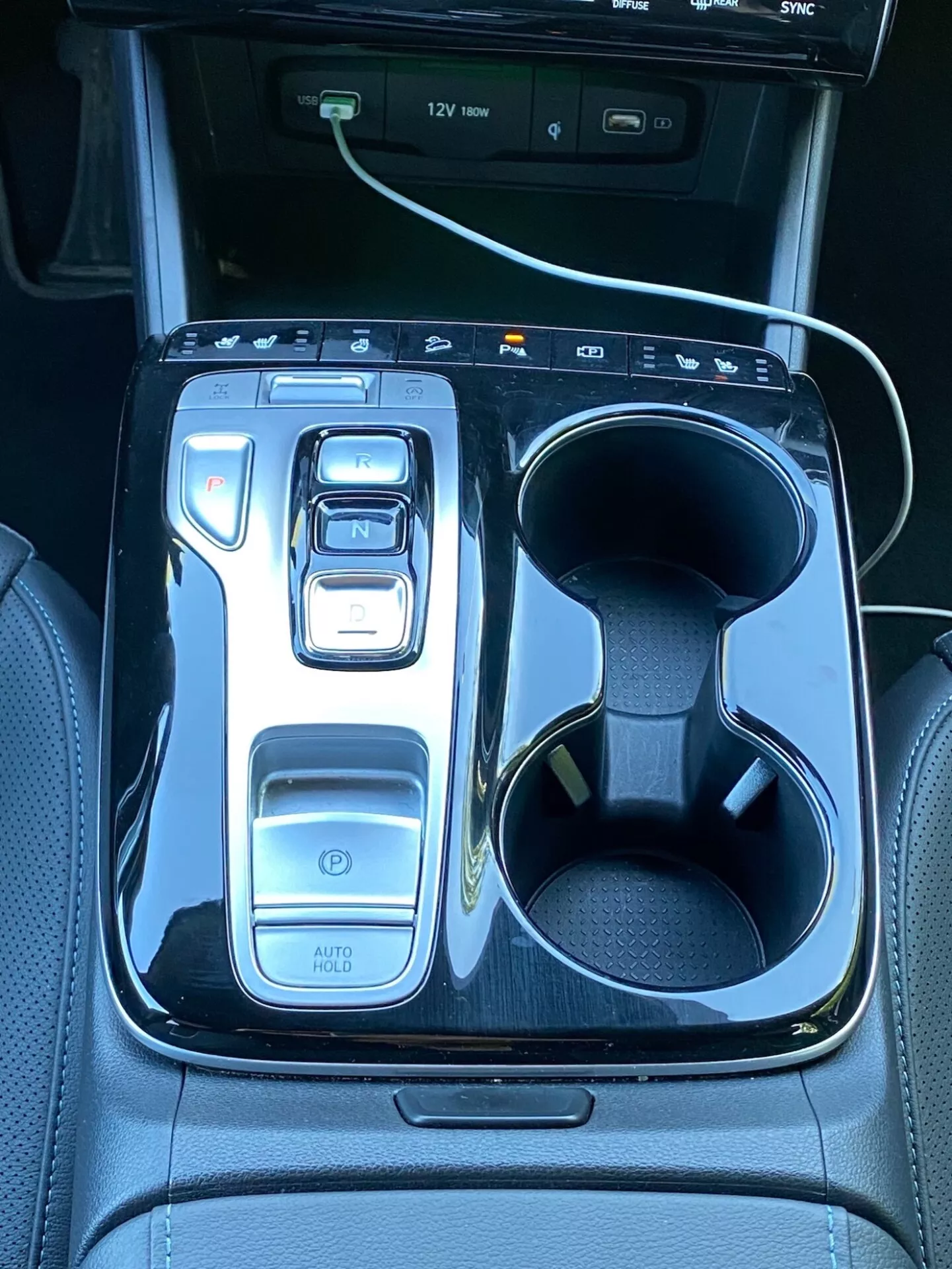 L'image montre la console centrale d'une Hyundai Tucson 2020, avec un sélecteur de vitesses électronique sur 'N' entouré d'une finition noire brillante. À gauche se trouvent des commandes comprenant un bouton d'alimentation, des sélecteurs de modes de conduite et la fonction 'AUTO HOLD'. À droite, il y a des porte-gobelets doubles avec un câble de charge blanc dans l'un des emplacements. Dans la partie supérieure, on aperçoit une partie de l'écran d'infodivertissement avec des indicateurs de port USB, mettant en valeur la technologie et le luxe du véhicule.