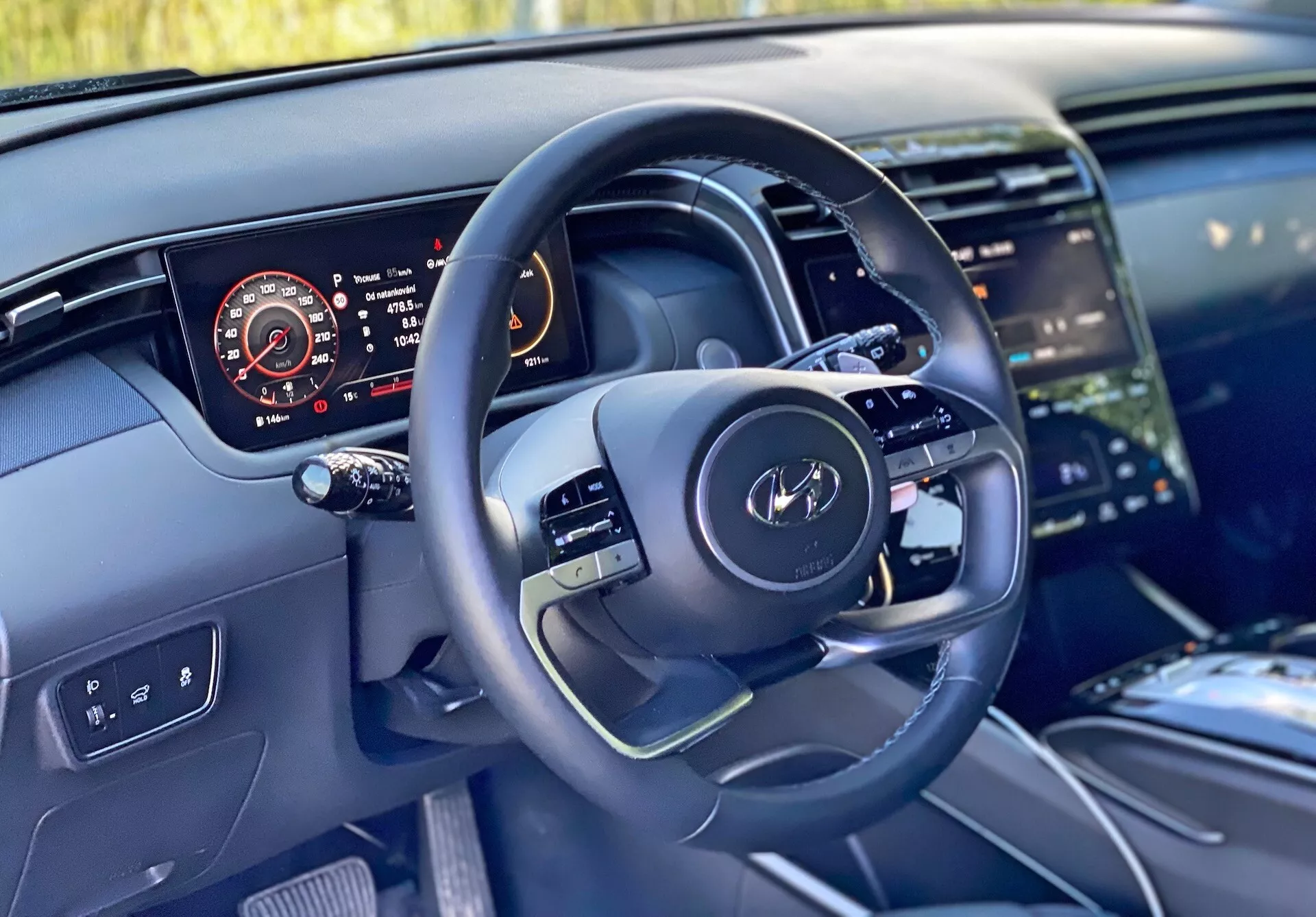 Vue intérieure d'une Hyundai Tucson 2020 depuis la perspective du conducteur mettant en avant le volant moderne avec commandes multifonctions et un tableau de bord numérique affichant le compteur de vitesse, le compte-tours, le niveau de carburant et la température du moteur. L'image met également en évidence une partie de la console centrale avec un grand système d'infodivertissement à écran tactile, un design de tableau de bord épuré avec éclairage d'ambiance et des matériaux de haute qualité suggérant des finitions de luxe.