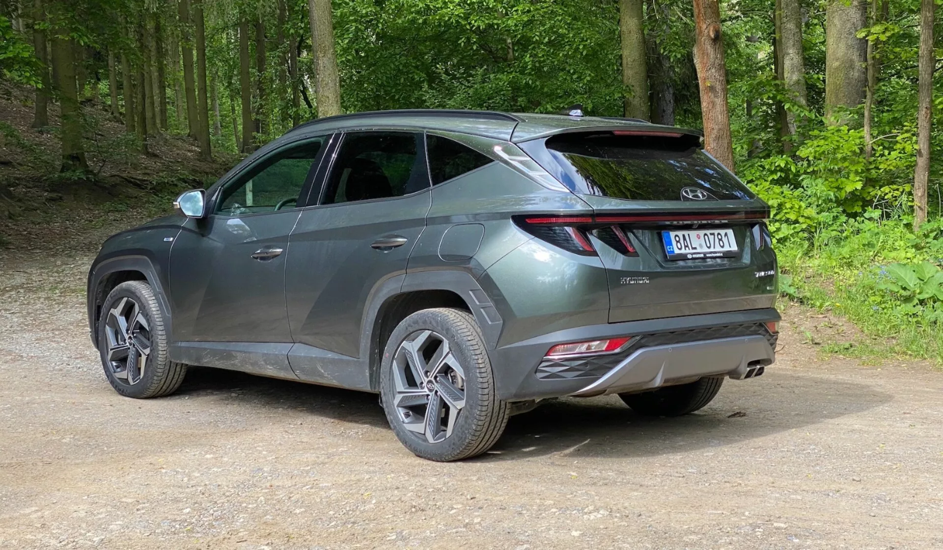 Un SUV moderne stationné sur un chemin non pavé dans une zone forestière, capturé d'une vue arrière trois quarts à une légère élévation. Le véhicule présente un design élégant et contemporain avec des lignes nettes, des feux arrière allongés qui s'enroulent de l'arrière vers les côtés, un spoiler visible et des réflecteurs sur le pare-chocs arrière, suggérant des fonctionnalités de sécurité avancées. Le véhicule gris métallique a des jantes en alliage à plusieurs branches et des pneus de route, avec une surface brillante indiquant qu'il est neuf ou bien entretenu. Autour de la voiture se trouve une forêt verte et luxuriante avec un sol en terre compactée et de la gravier lâche.