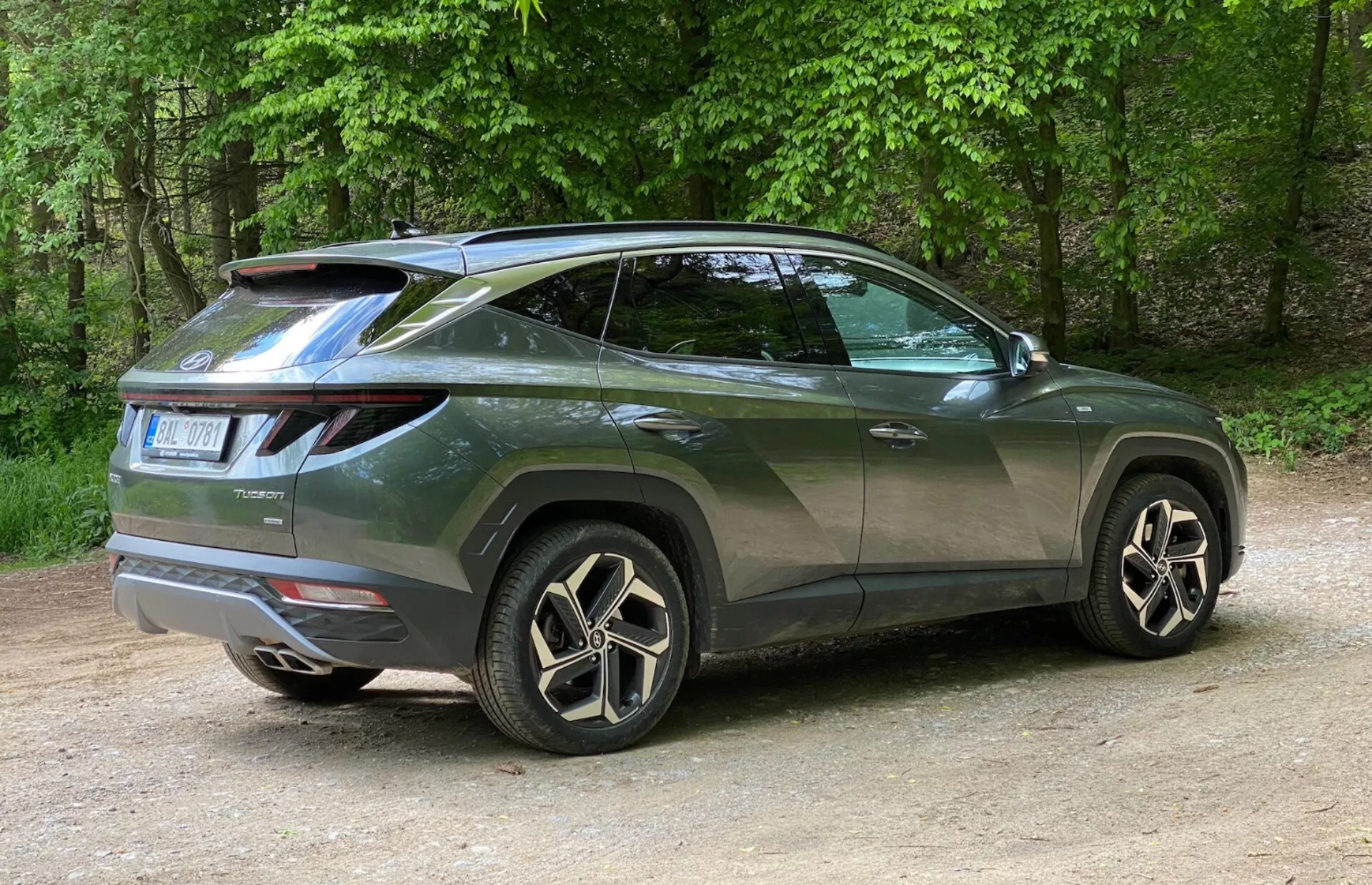 Vue de trois quarts arrière d'un SUV moderne Hyundai Tucson 2020 mettant en avant son profil latéral et son design arrière. Le véhicule est positionné à gauche avec la photo prise de droite, mettant en avant l'angle arrière droit. Les feux arrière à LED distinctifs s'enroulent de l'arrière vers les côtés et un spoiler monté sur le toit ajoute une touche sportive. La ligne de ceinture ascendante, le toit incliné, les vitres teintées et les poignées de porte de la couleur de la carrosserie accentuent son apparence de coupé. Les grandes jantes en alliage avec un design à multiples rayons et les pneus à profil bas rehaussent son aspect luxueux. Les petits détails comme l'essuie-glace arrière et les moulures des passages de roues signifient praticité et robustesse. La voiture est placée sur du gravier dans un cadre naturel avec des arbres en arrière-plan.