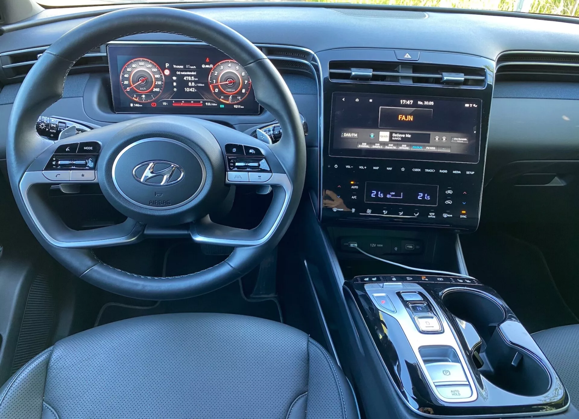 Vue intérieure de la Hyundai Tucson 2020 depuis le siège du conducteur, montrant le volant avec le logo Hyundai et les commandes montées pour audio, téléphone et paramètres du régulateur de vitesse. Le tableau de bord dispose d'un panneau d'instruments numérique avec compteur de vitesse et tachymètre accentués en rouge. La console centrale comprend un système d'infodivertissement avec écran tactile affichant les commandes de climatisation, des boutons physiques et des cadrans pour le contrôle de la température, et un levier de vitesses automatique avec sélecteur électronique. Un socle de recharge sans fil potentiel est visible, avec un intérieur en tissu sombre et un habitacle bien éclairé.