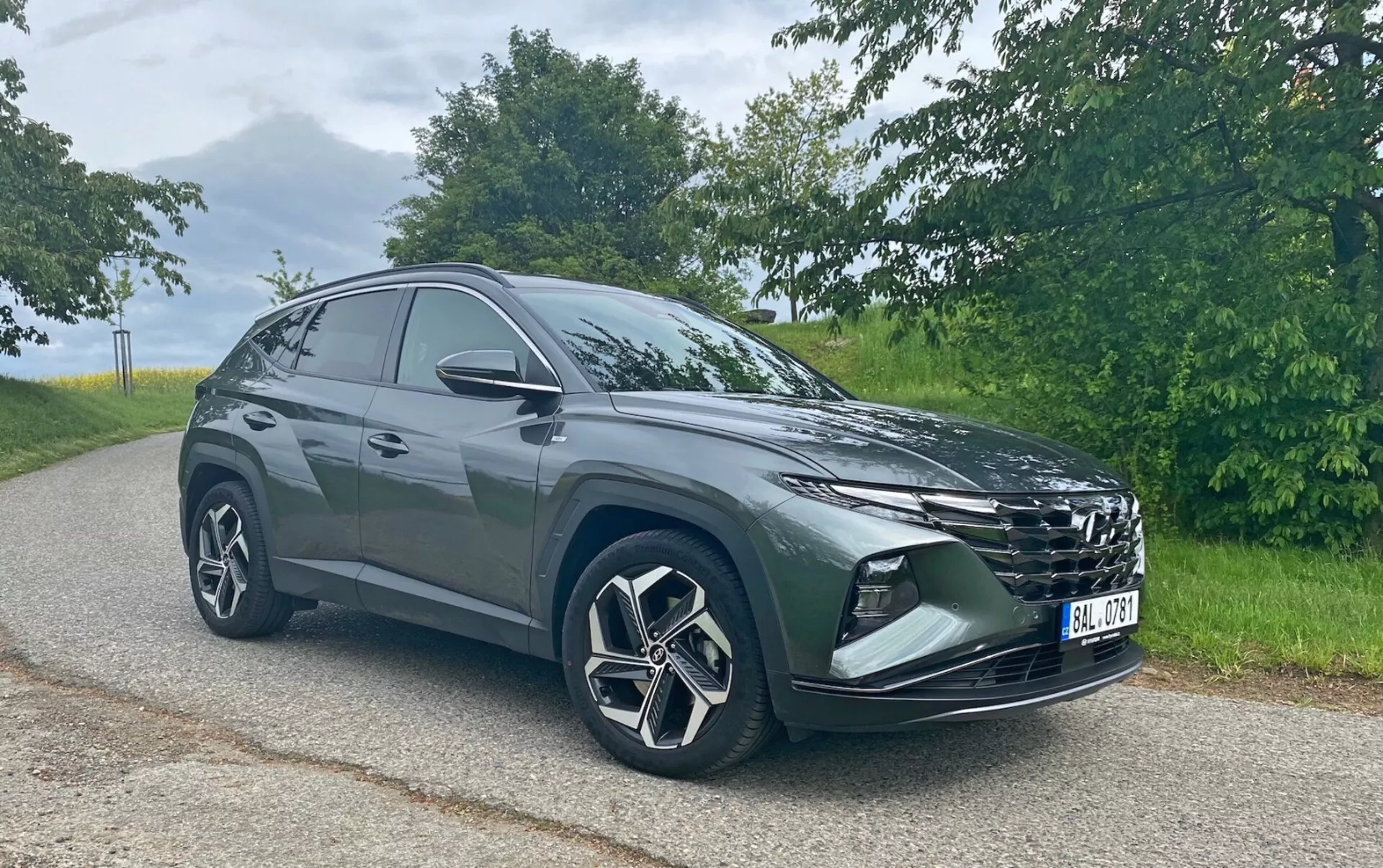 L'image montre un SUV Hyundai Tucson 1.6 T-GDi Pure moderne garé sur une route asphaltée avec un paysage naturel en arrière-plan. Photographié sous un angle trois-quarts avant, on peut voir la calandre proéminente, les phares élégants, le pare-chocs avant avec des phares antibrouillard ou des prises d'air, les grandes roues en alliage avec un design à multi-rayons et les rétroviseurs latéraux. La silhouette du SUV montre une ligne de toit inclinée vers l'arrière, indicative d'un design sportif et aérodynamique. Capturée par une lumière douce en journée, la photo met en valeur la couleur et les détails du véhicule sans ombres fortes.