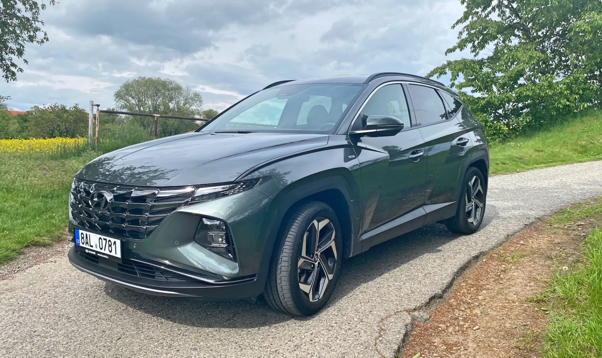 Hyundai Tucson 2020 1.6 T-GDI Pure SUV, vu de trois quarts avant sur une route pavée avec des arbres et de l'herbe en arrière-plan. L'image montre la calandre du véhicule, les phares LED aiguisés, les jantes en alliage, les rétroviseurs latéraux avec clignotants intégrés et une peinture métallisée foncée. Les portes avant et arrière côté conducteur sont visibles. Photo prise en journée par temps couvert.