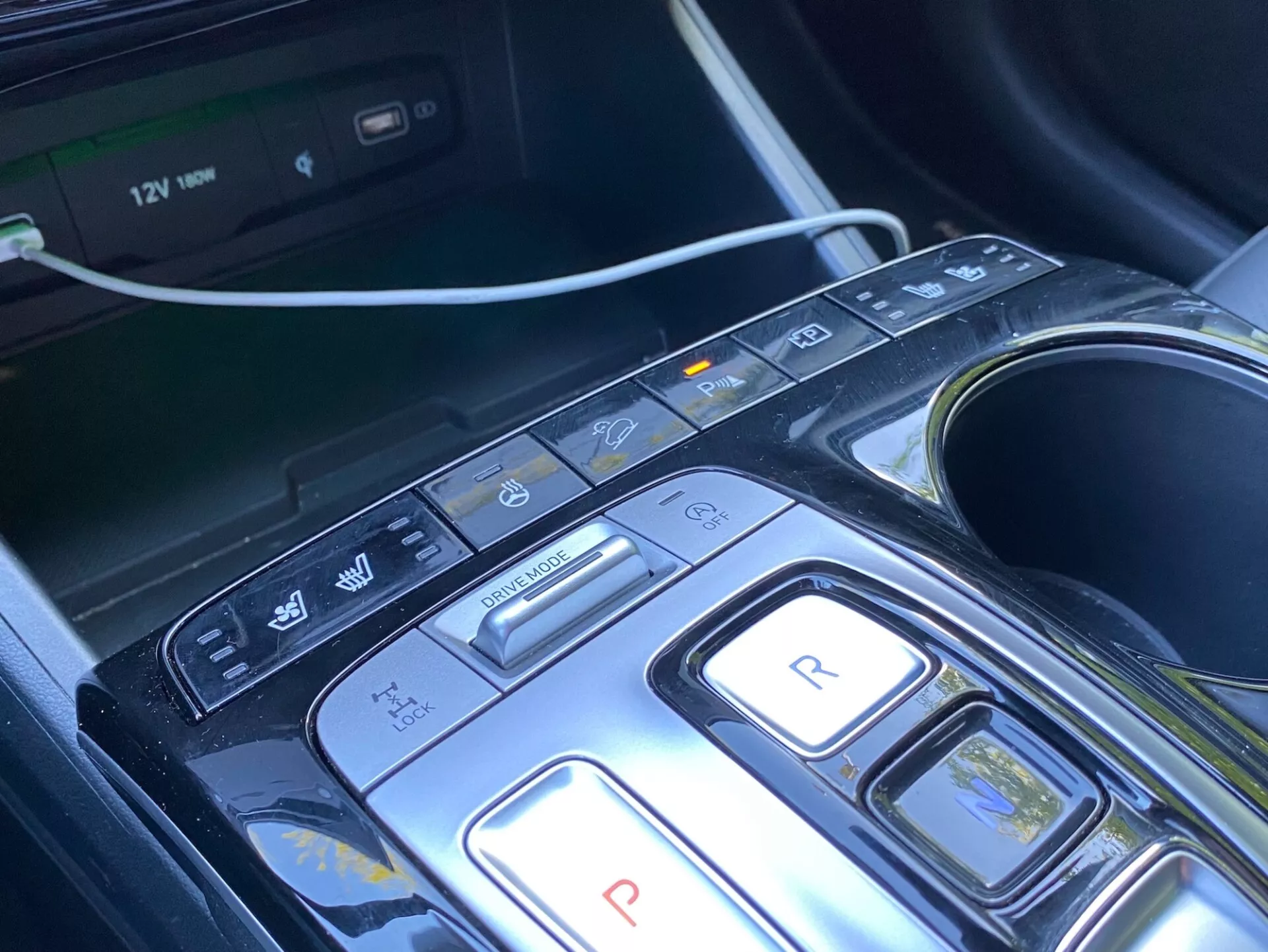 Gros plan sur la console centrale et la zone de sélection de vitesse d'un Hyundai Tucson 2020 1.6 T-GDi Pure, montrant un design moderne avec des boutons pour le stationnement, la marche arrière, le point mort, la conduite et les modes de frein/ régénératif, avec des boutons supplémentaires pour l'arrêt/démarrage automatique, les capteurs de stationnement/le stationnement automatisé et l'avertissement de maintien de voie/départ de voie. Écran du système multimédia visible, porte-gobelets et prise de courant 12V indiquant confort, commodité et technologies avancées d'assistance au conducteur (ADAS). La console a un design élégant avec du matériau noir brillant et des accents métalliques, suggérant une allure de luxe.