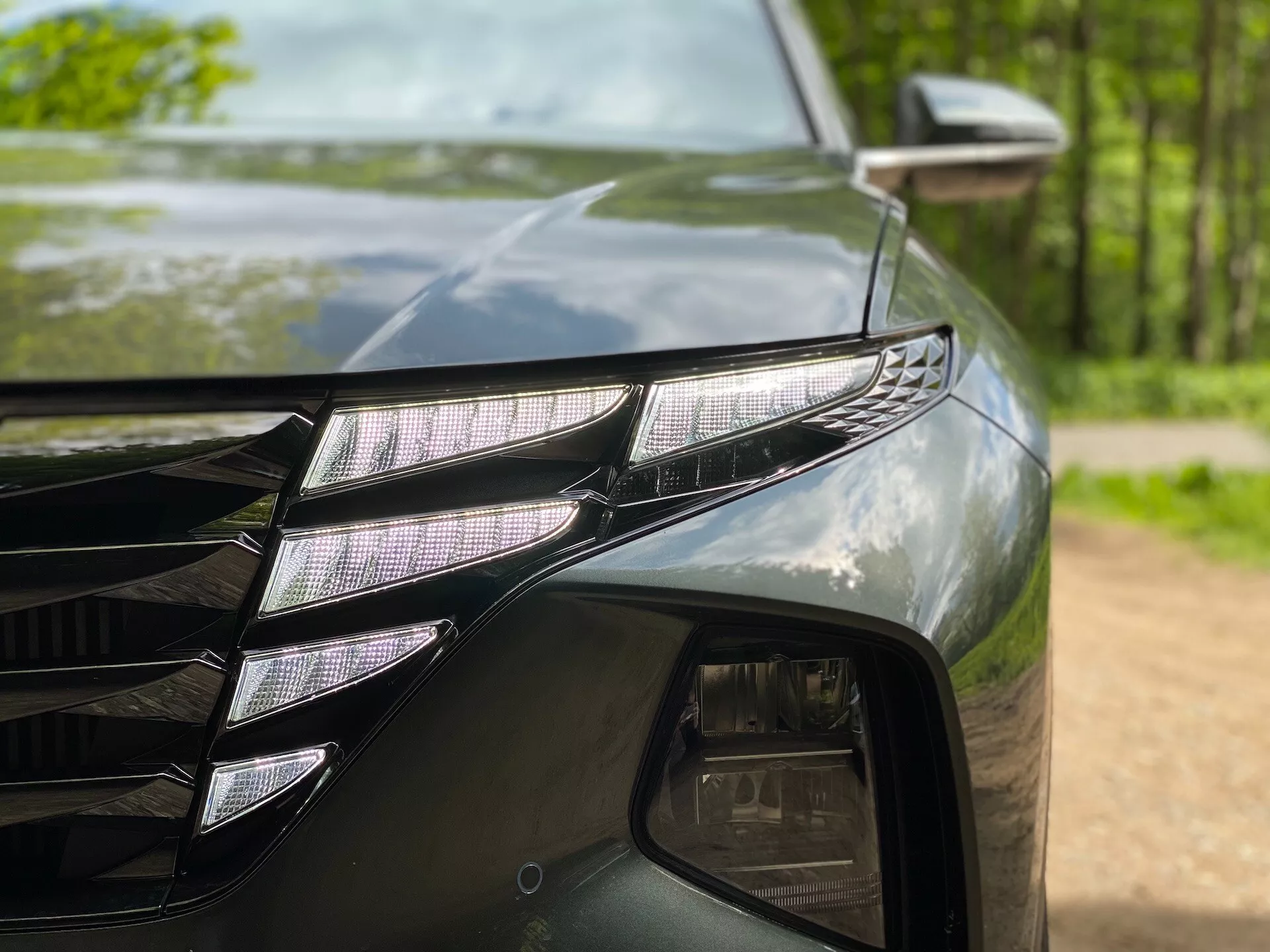 Gros plan sur le côté avant gauche d'une Hyundai Tucson 2020 (Hyundai-Tucson-1-6-T-GDi-Pure), mettant en avant un ensemble de phares LED avec des projecteurs LED individuels et une bande LED DRL distinctive. La peinture sombre et brillante reflète les arbres environnants. On peut également observer une partie de la calandre aux lignes angulaires et le rétroviseur latéral avec un indicateur de direction.
