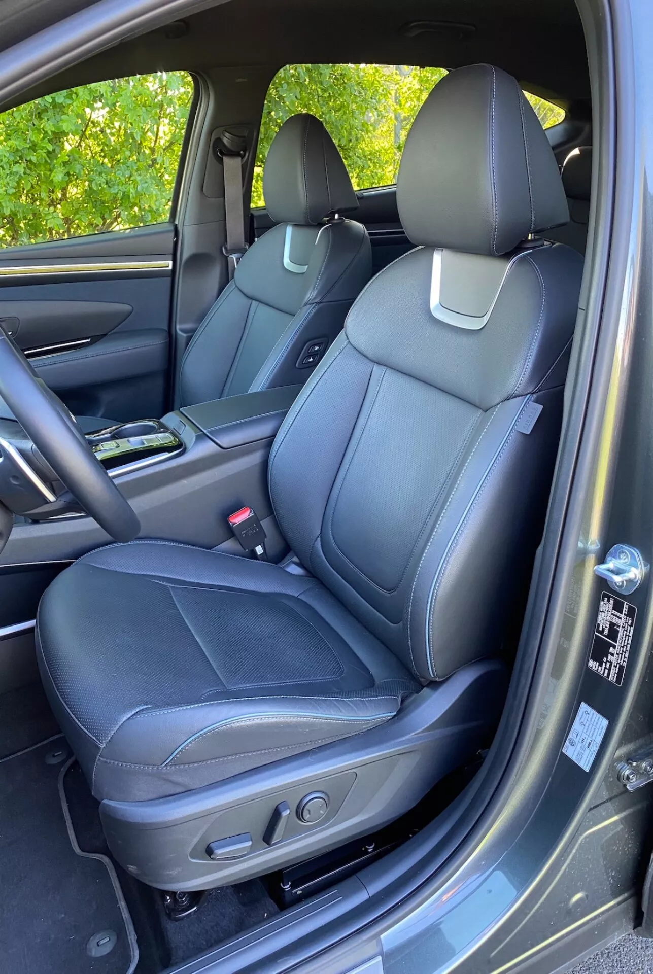 Intérieur du Hyundai Tucson 2020 1.6 T-GDI Pure montrant le siège passager avant avec une sellerie en cuir noir, des coutures détaillées et des commandes de réglage électrique. Console centrale visible avec porte-gobelets, panneau de porte avec matériaux assortis, poignée, compartiment de rangement et grille de haut-parleur suggérant un système audio avancé. Tableau de bord et boîte à gants partiellement visibles. Tapis de sol en moquette et léger reflet de lumière du soleil sur le panneau de porte.