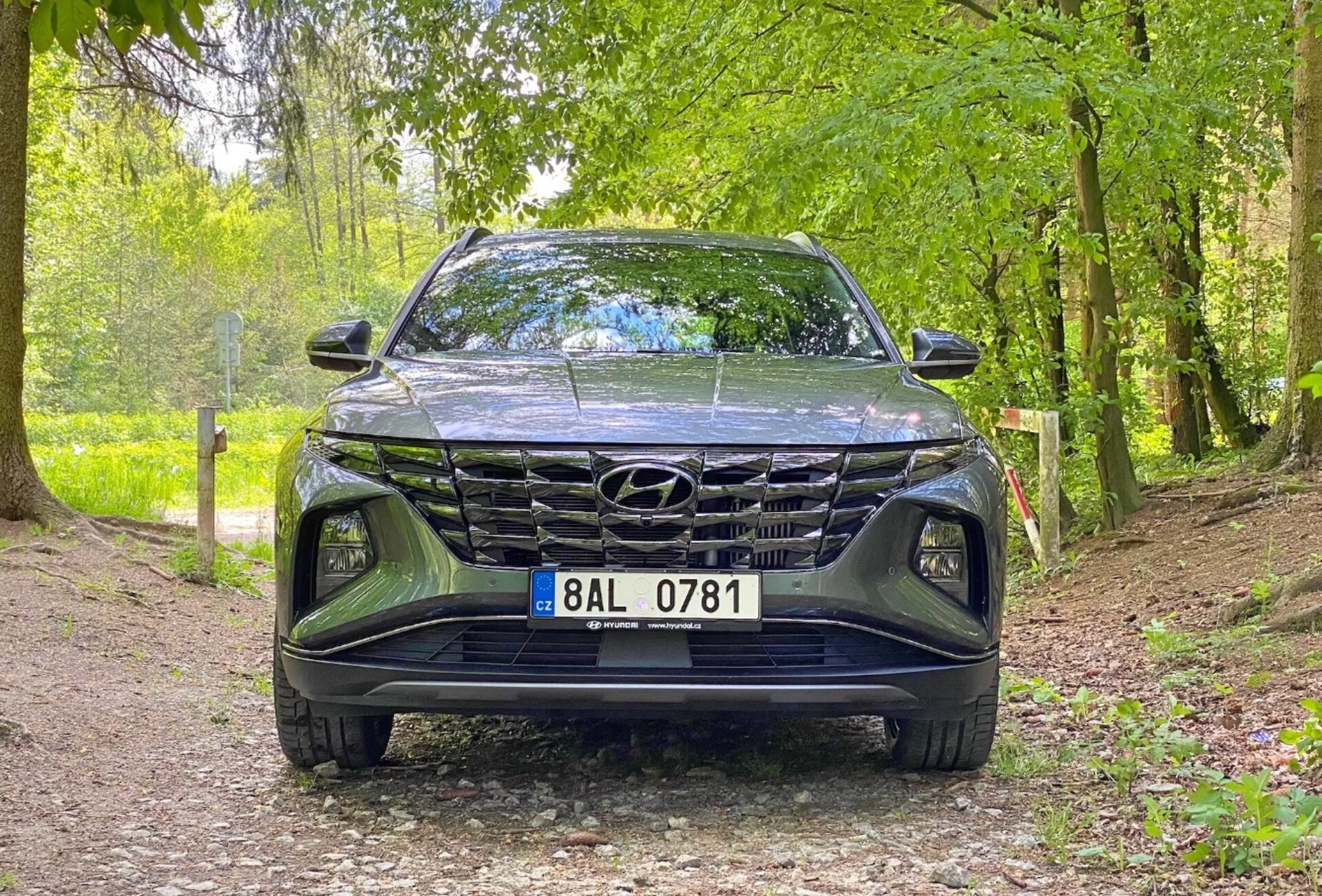 Vue frontale du véhicule Hyundai Tucson 1.6 T-GDi Pure 2020, présentant une prise de vue en contre-plongée centrée sur la calandre, le pare-chocs avant et la zone du capot. La voiture est garée sur un chemin de gravier dans un environnement naturel, suggérant son adéquation pour les environnements extérieurs. Les caractéristiques clés incluent un design de calandre proéminent, des phares à LED élégants et le badge Hyundai visible.