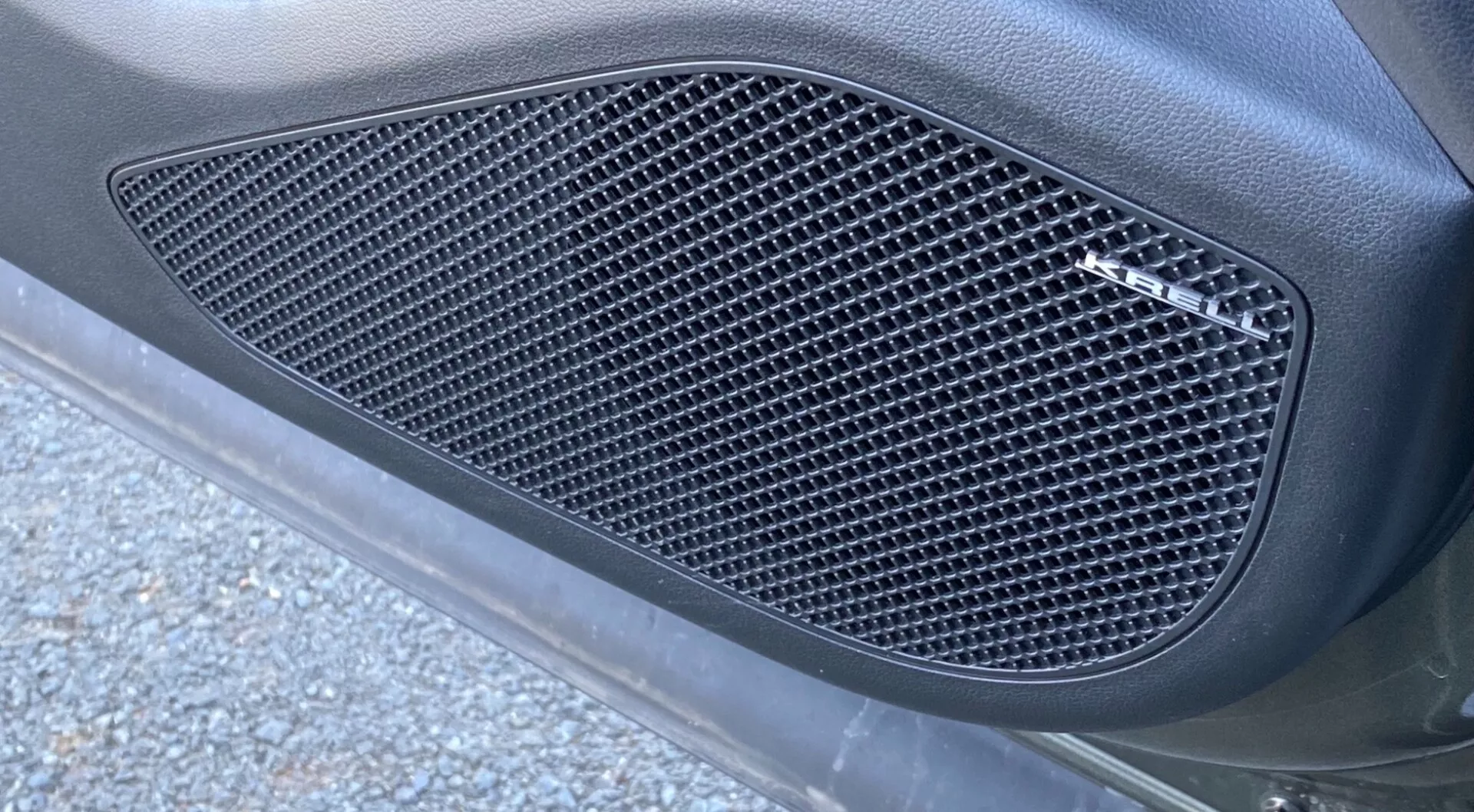 Vue de côté en gros plan du Hyundai Tucson 2020 1.6 T-GDI Pure, se concentrant sur une grille latérale avec un motif en treillis de découpes en forme de diamant. La grille, encastrée dans un contour moulé, est intégrée dans le panneau de carrosserie de la voiture avec une finition mate, entourée d'une surface lisse et incurvée. L'image, prise sous un angle oblique de bas en haut, montre un éclairage naturel extérieur avec des morceaux de bitume et des débris de route, améliorant l'apparence élégante et axée sur la performance.
