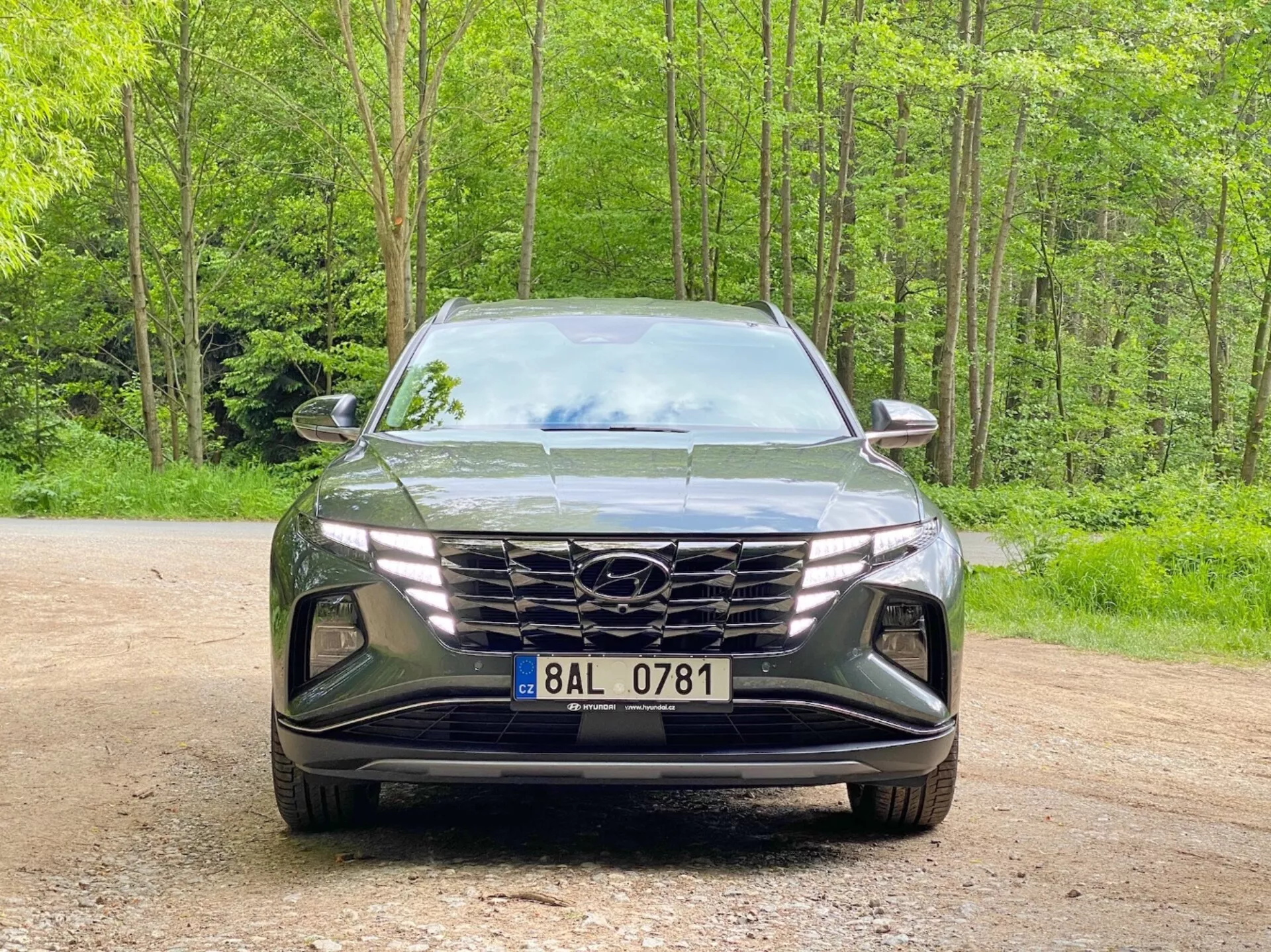 Vue de face d'un Hyundai Tucson 2020 (Hyundai Tucson 1.6 T-GDI Pure) SUV moderne garé sur un chemin de gravier avec un fond forestier. L'image capture le style élégant et agressif avec une grande calandre géométrique mettant en avant le logo du fabricant, des phares LED angulaires étroits et un pare-chocs avant sculpté avec des prises d'air et des logements pour les phares antibrouillard, soulignant l'aérodynamique avancée et la technologie d'éclairage.