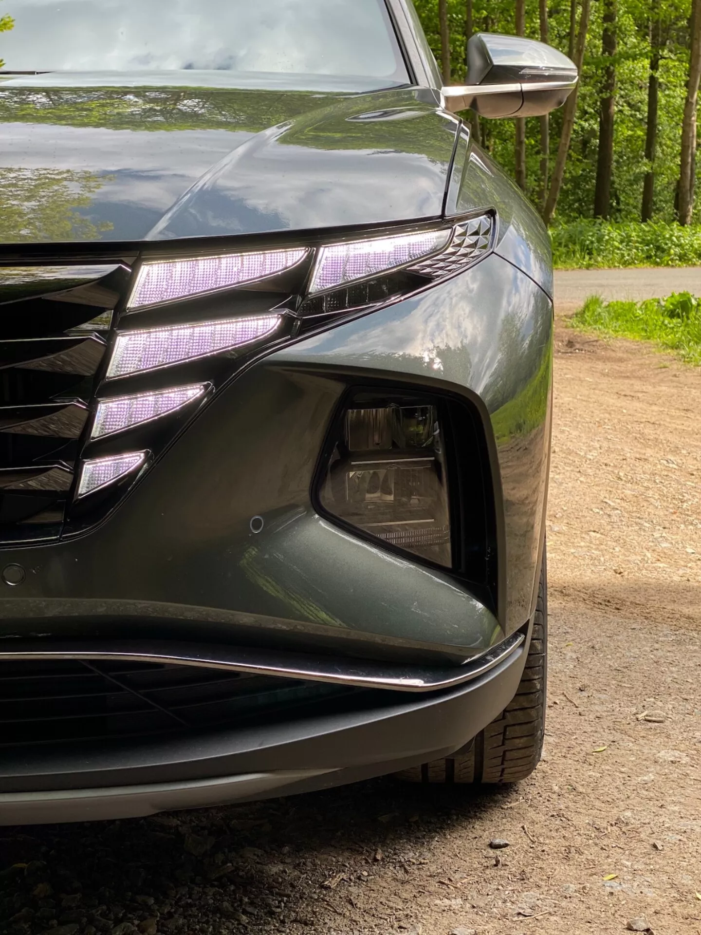 Vue rapprochée de l'angle avant gauche d'une Hyundai Tucson 2020 1.6 T-GDI Pure, mettant en avant le phare avec feux de jour à LED et lentille de type projecteur, une partie de la calandre avant avec des lignes horizontales et des accents chromés, la section de prise d'air avec logement pour phares antibrouillard, le pare-chocs avant sculpté avec des lignes fluides et le clignotant intégré dans le rétroviseur extérieur. La peinture sombre et brillante met en valeur les courbes aérodynamiques de la voiture.