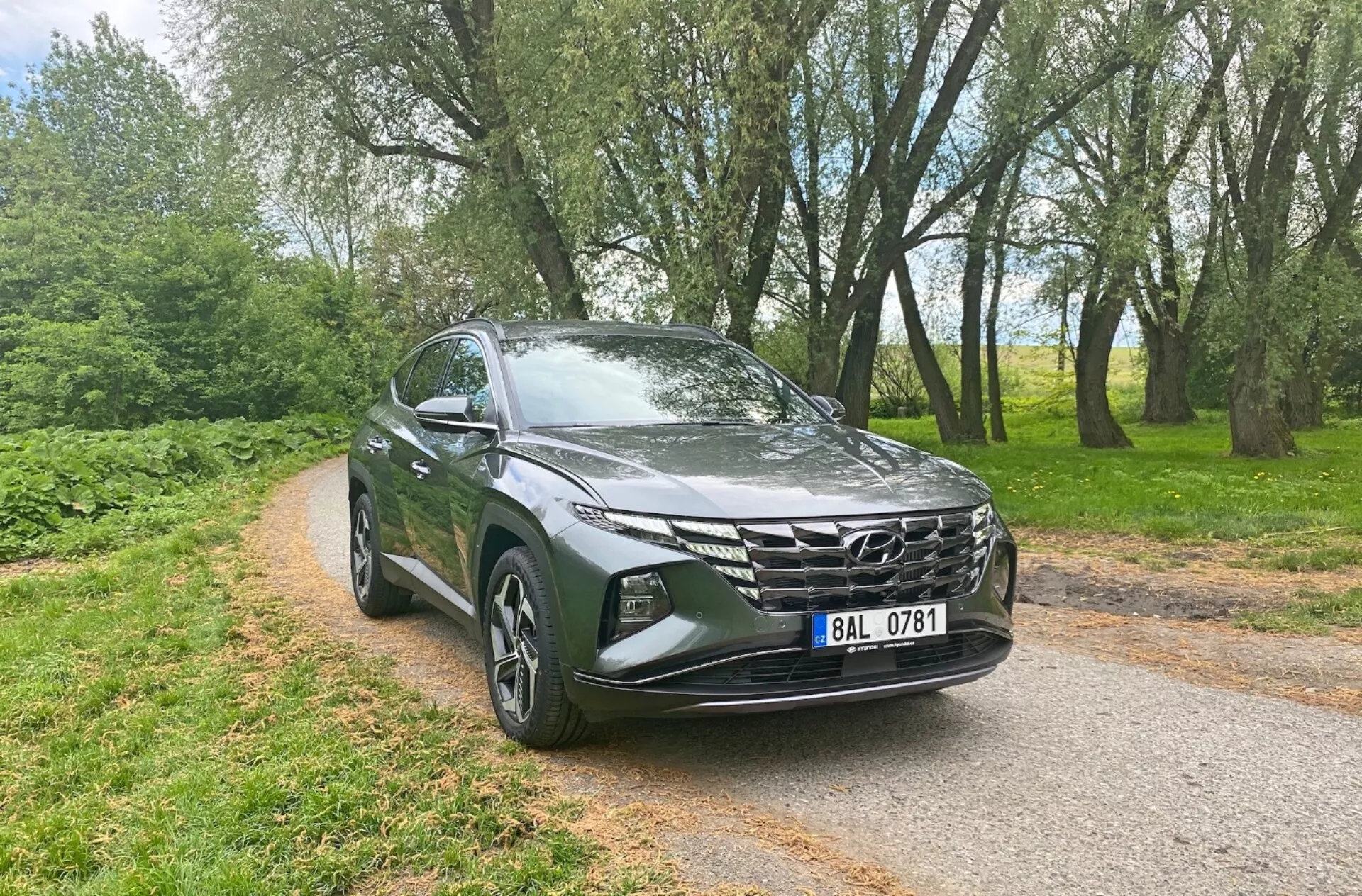 Vue avant trois quarts d'un Hyundai Tucson 1.6 T-GDi Pure 2020, montrant des caractéristiques modernes de SUV comme des phares LED élégants, une calandre avant géométrique proéminente, un capot aérodynamique sculpté, des jantes en alliage, des rétroviseurs latéraux avec indicateurs de direction intégrés et une peinture métallisée, sur une route étroite pavée bordée d'arbres en arrière-plan, soulignant sa polyvalence et son attrait pour les environnements urbains et ruraux.