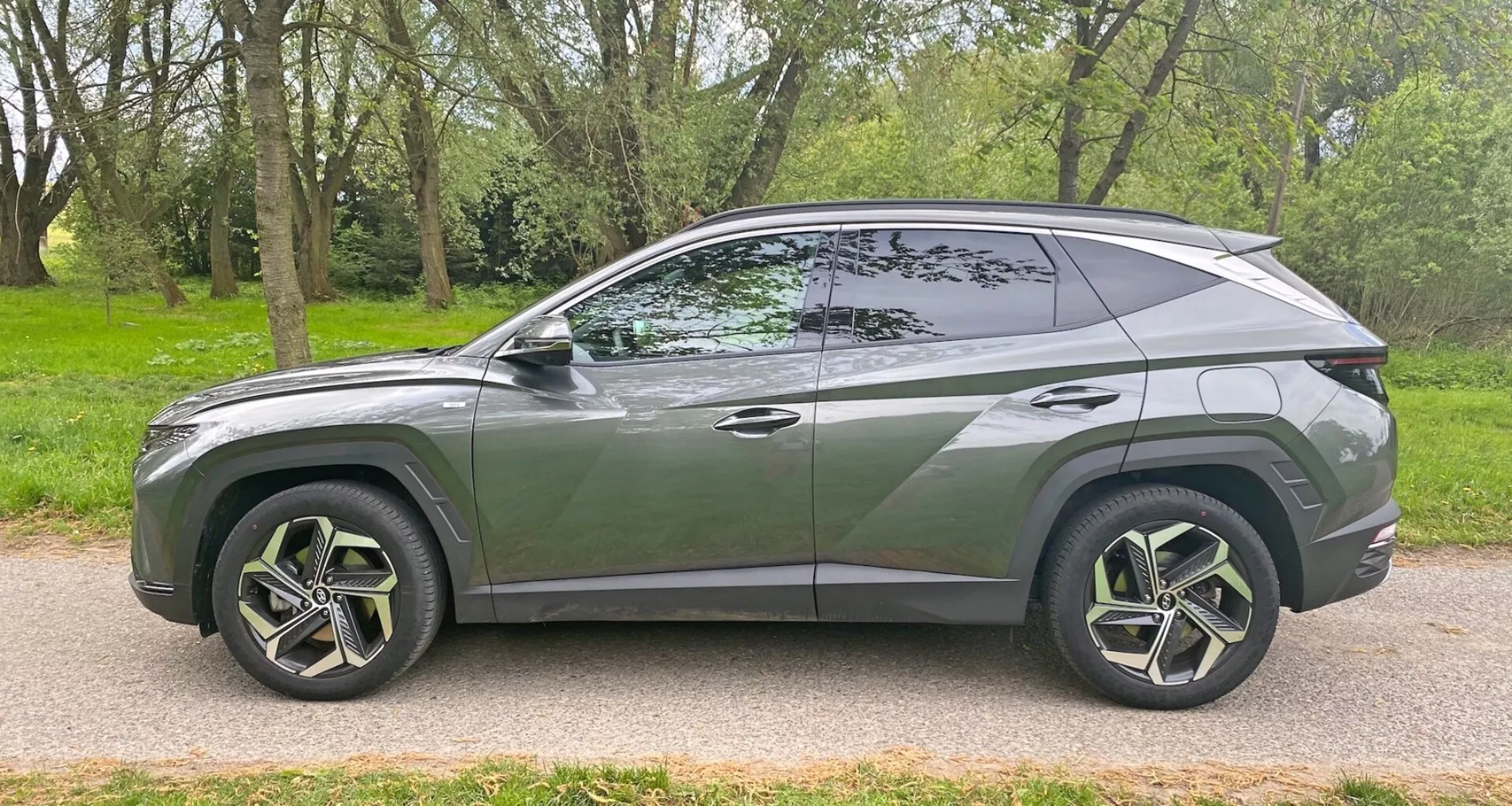 Profil latéral du moderne SUV Hyundai Tucson 1.6 T-GDI Pure 2020 garé sur une route pavée à côté d'une zone herbeuse avec des arbres. Montrant son design aérodynamique et élégant avec une ligne de toit descendante, une ligne de carrosserie distinctive, de grandes roues en alliage à plusieurs rayons avec des pneus à profil bas, des fenêtres teintées avec un pilier B visible, des poignées de porte affleurantes, des rétroviseurs body colorés, et une garniture chromée le long du bord de la fenêtre. Le véhicule capture un style contemporain et des caractéristiques avancées potentielles sous un angle latéral.