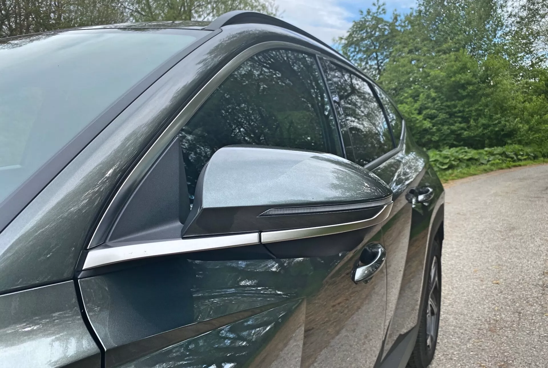 Vue latérale en gros plan de la Hyundai Tucson 2020 1.6 T-GDI Pure mettant en avant son design moderne et ses fonctionnalités technologiques, centrée sur le côté conducteur. La photographie en contre-plongée met en évidence le rétroviseur latéral élégant et aérodynamique avec indicateurs de signalisation intégrés et accents noirs, suggérant des fonctionnalités avancées. La poignée de porte avant du conducteur, affleurante, indique un système d'entrée sans clé, contribuant à l'efficacité aérodynamique du véhicule. Les portes de couleur carrosserie brillante et les garnitures de fenêtre en chrome évoquent un pack finition haut de gamme. Les éléments de fond comme les arbres et le ciel suggèrent un environnement extérieur.