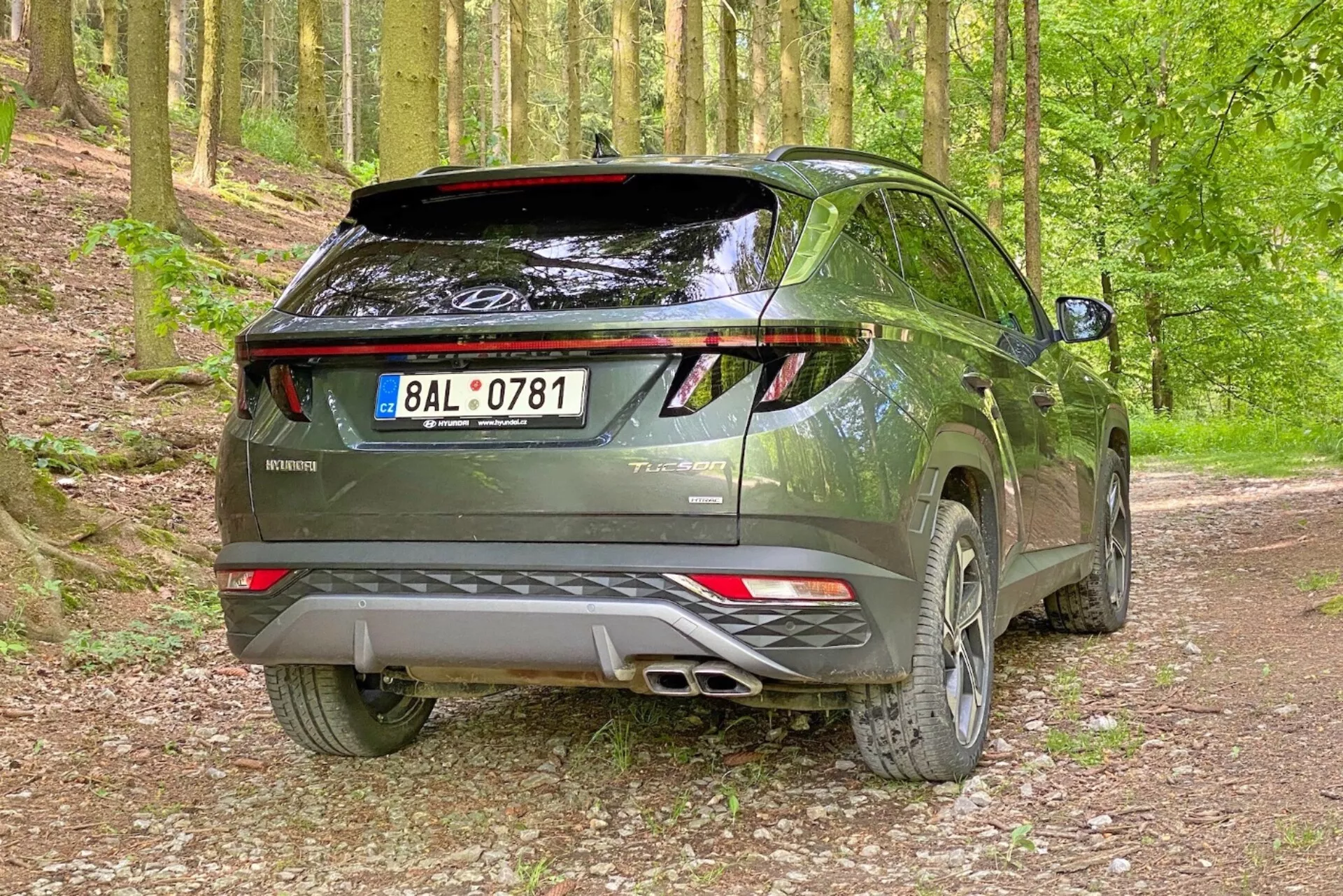 Image d'un SUV moderne Hyundai Tucson 2020 dans un cadre forestier sur un chemin de gravier, capturée sous un angle arrière trois quarts. La photo met en évidence le côté droit et le profil arrière du véhicule avec un hayon arrière visible, des feux arrière à LED, un système d'échappement double, un becquet arrière et des jantes en alliage.