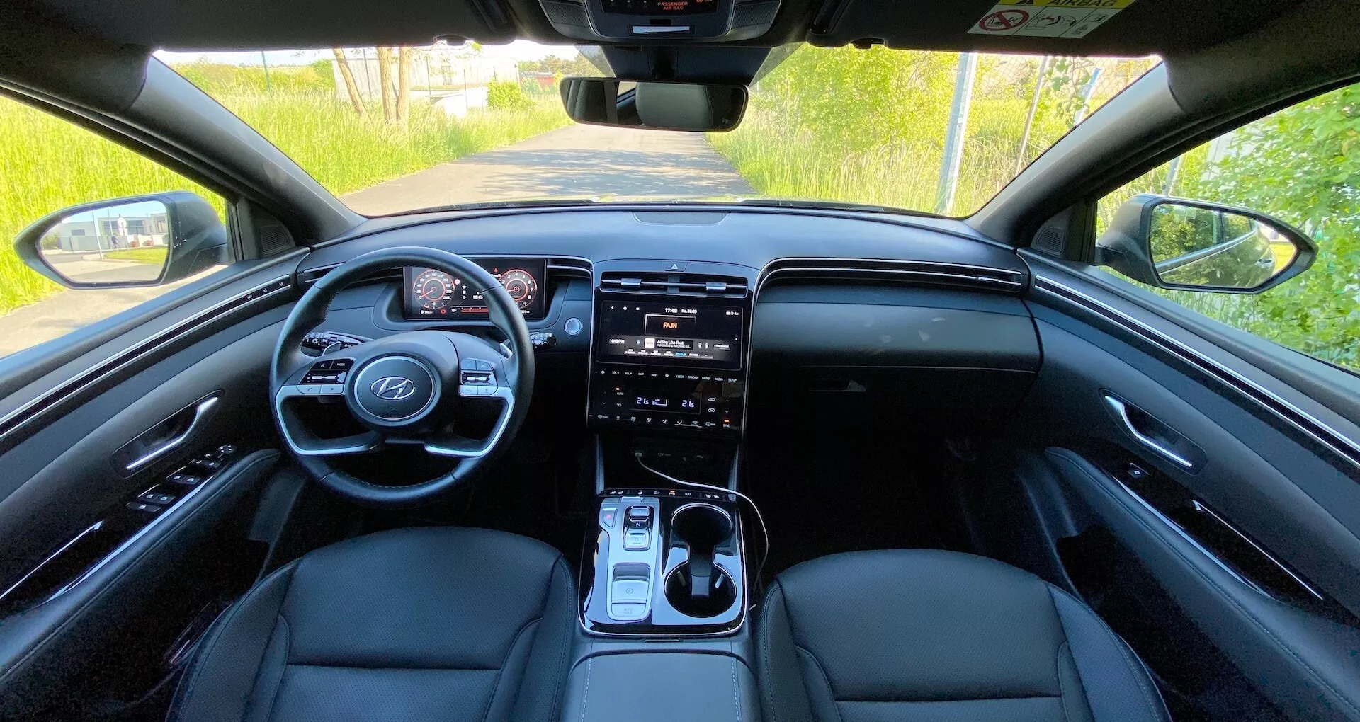 Vue intérieure de la Hyundai Tucson 2020 depuis la banquette arrière montrant le tableau de bord avant, volant avec commandes intégrées, tableau de bord numérique, grand système d'infodivertissement à écran tactile, boutons de contrôle du climat, levier de vitesse et sièges avant en cuir sous l'éclairage naturel de jour.