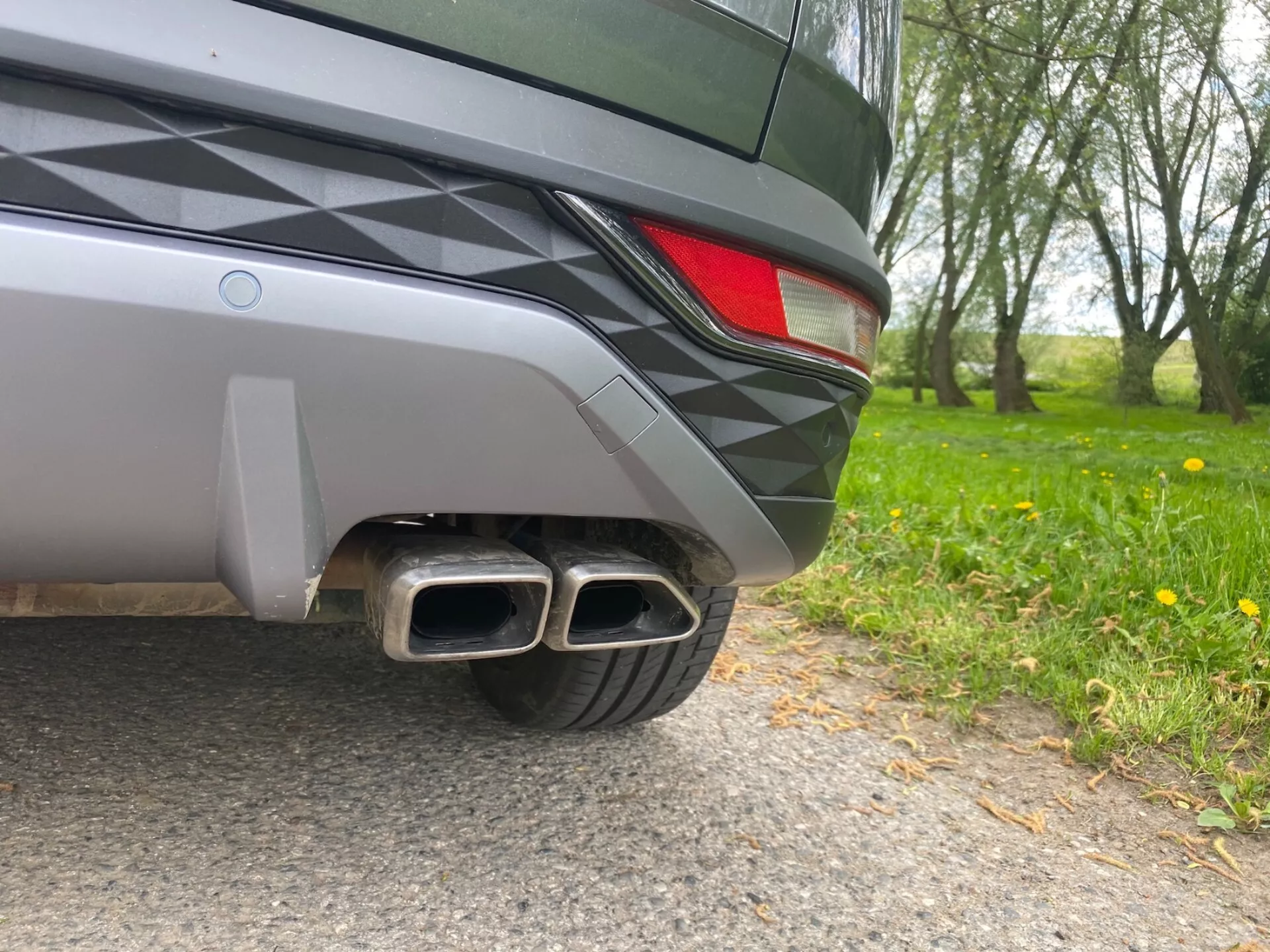 Image en gros plan de la section arrière d'une Hyundai Tucson 2020, prise à un angle au niveau du sol. Les détails visibles comprennent un pare-chocs arrière bicolore, une partie du feu arrière à LED et des doubles tuyaux d'échappement métalliques avec des embouts rectangulaires, suggérant une finition sportive. Le design élégant du véhicule et la peinture métallisée reflètent les environs naturels.
