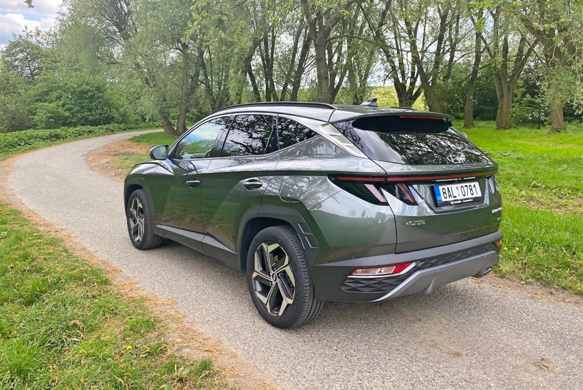 L'image montre un crossover SUV moderne sous un angle arrière trois quarts, mettant en évidence le design aérodynamique élégant et la ligne de toit inclinée. Le véhicule vert foncé présente une porte arrière de style liftback, un aileron sportif et de grandes roues en alliage avec un design à plusieurs branches. Les éléments visibles incluent des feux arrière à LED modernes, des sorties d'échappement doubles et un essuie-glace arrière. Le SUV est garé sur un chemin de gravier entouré de verdure, suggérant un environnement de parc ou rural.
