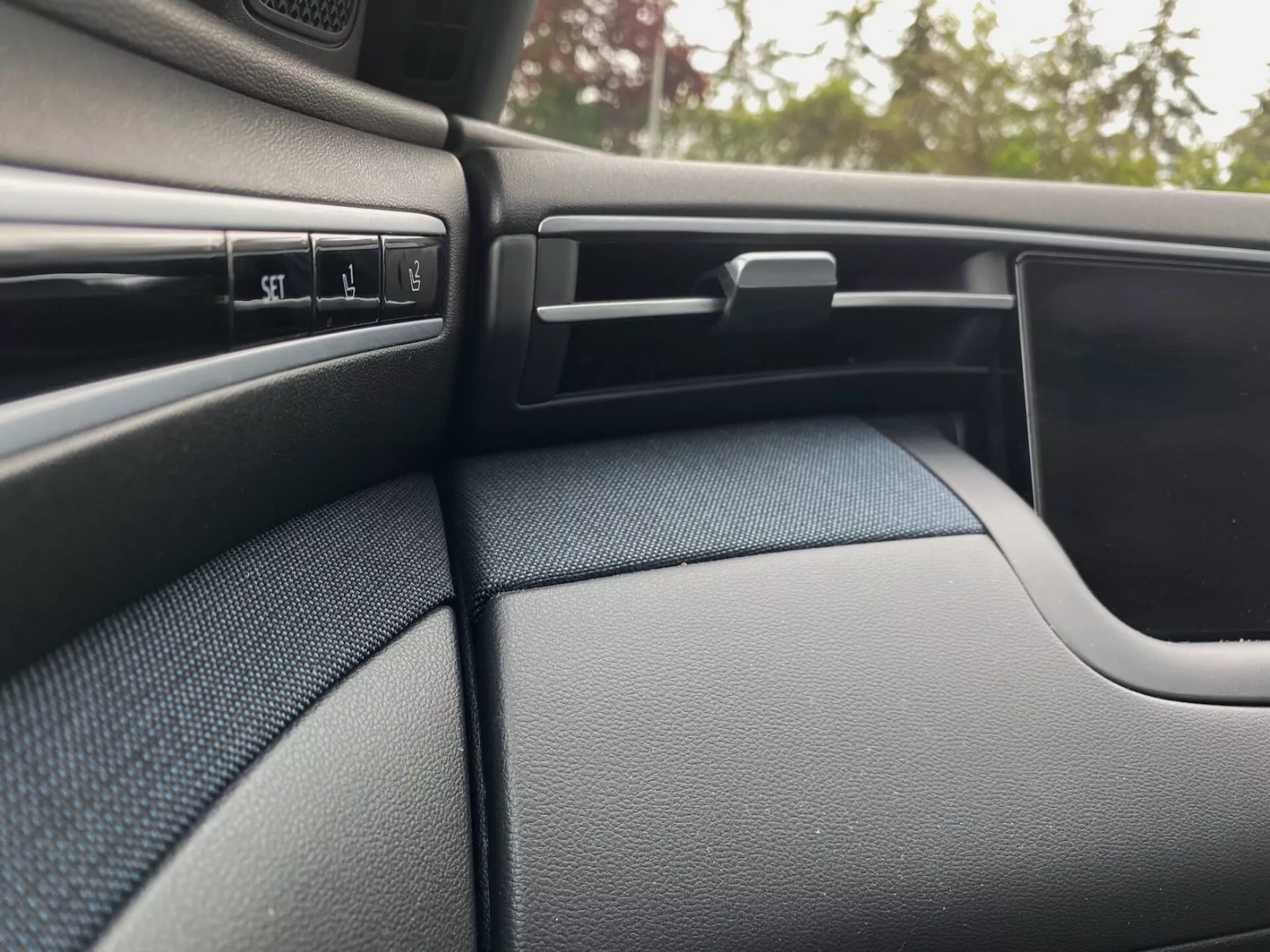 Détail de l'intérieur du Hyundai Tucson 2020, montrant le tableau de bord texturé de couleur sombre du côté passager et une partie du panneau de porte. La scène met en évidence plusieurs boutons de commande, une bouche d'aération réglable avec des lamelles horizontales, une boîte à gants verrouillable et le design incurvé du panneau de porte qui se fond harmonieusement dans le tableau de bord.