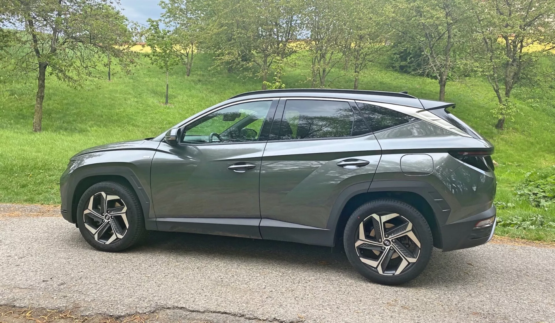 Vue de profil latéral d'un SUV Hyundai Tucson 1.6 T-GDI Pure 2020 en plein jour. Le véhicule est positionné dans une zone herbeuse avec des arbres en arrière-plan, suggérant un environnement rural ou suburbain. Les caractéristiques de design moderne incluent des phares LED, des clignotants intégrés aux rétroviseurs latéraux, des vitres teintées, des jantes en alliage aérodynamiques et une ligne de toit inclinée rappelant un coupé. La carrosserie élégante avec des lignes de caractère et des poignées de porte affleurantes suggèrent un système d'entrée sans clé avancé, tandis que le spoiler arrière indique des améliorations aérodynamiques. La voiture est centrée dans le cadre, offrant une vue claire et dégagée depuis une perspective de hauteur moyenne.