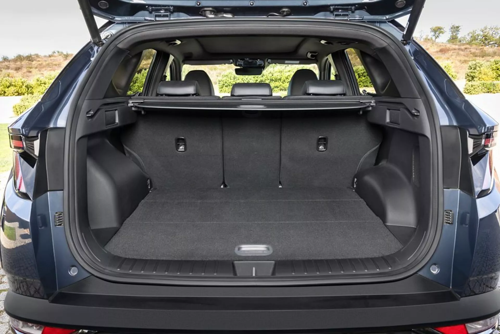 Zone de chargement arrière de Hyundai Tucson 2020 avec le coffre ouvert, montrant le plancher de chargement moquetté, des points d'ancrage, des compartiments de rangement, une prise 12V, des sièges arrière rabattables, une couverture de chargement rétractable, des parois latérales en plastique durable, et un aperçu du pare-chocs arrière, dans un cadre extérieur.