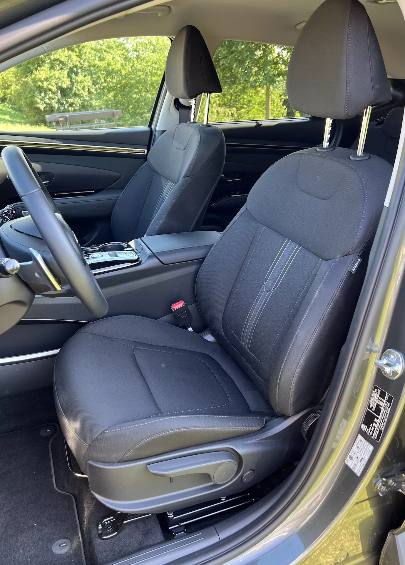 Vue intérieure du Hyundai Tucson 2020 centrée sur le siège conducteur avec une sellerie en cuir matelassé, appuie-tête intégré et commandes sur le panneau de porte pour les vitres et les rétroviseurs électriques, prise d'un angle légèrement élevé et oblique montrant les commandes d'ajustement du siège, une partie du volant, le sol tapissé et un aperçu de l'éclairage extérieur