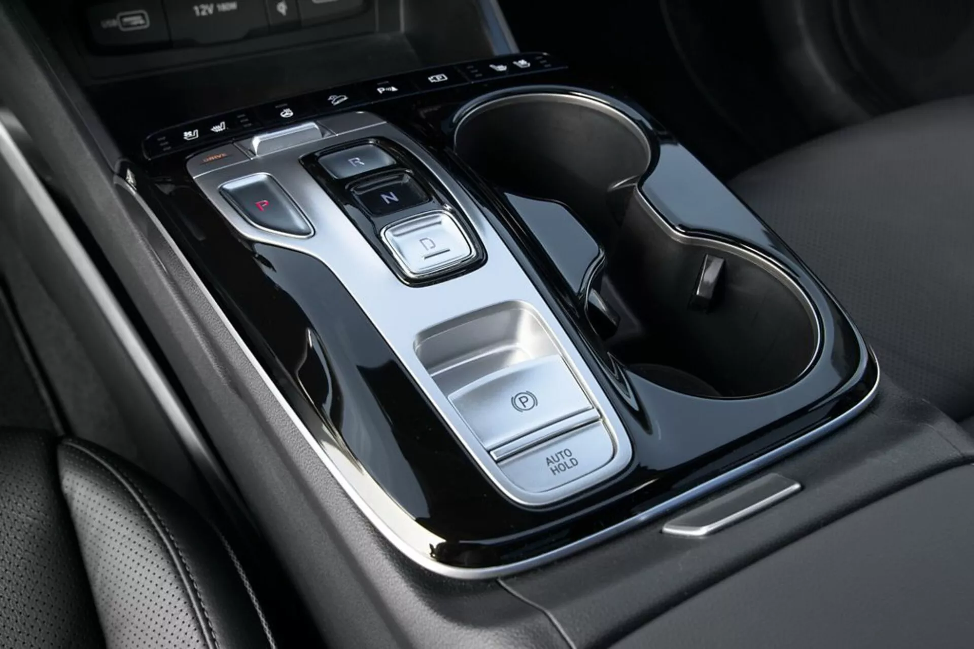 Gros plan sur la console centrale de la Hyundai Tucson 2020 montrant le levier de vitesse automatique, les boutons de commande, la fonction Auto Start/Stop, l'interrupteur des feux de détresse, et les porte-gobelets doubles avec des accents chromés, vu d'en haut avec une légère inclinaison.