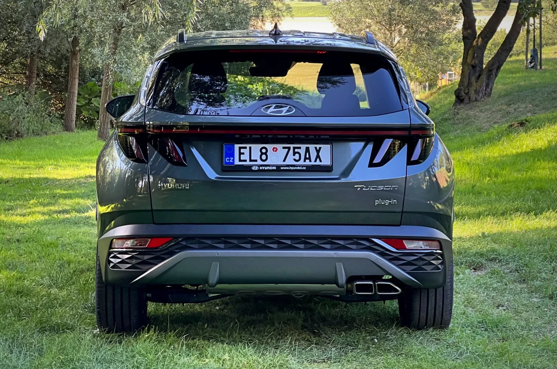 Vue arrière du SUV Hyundai Tucson 2020 sur pente herbeuse avec arrière-plan forestier, affichant logo sur hayon, badges Tucson et Hybrid, signe Plug-in, lumières réfléchissantes, feux arrière dynamiques, essuie-glace arrière, feu de stop haut placé, embouts d'échappement doubles, plaques de protection argentées et badge AWD.