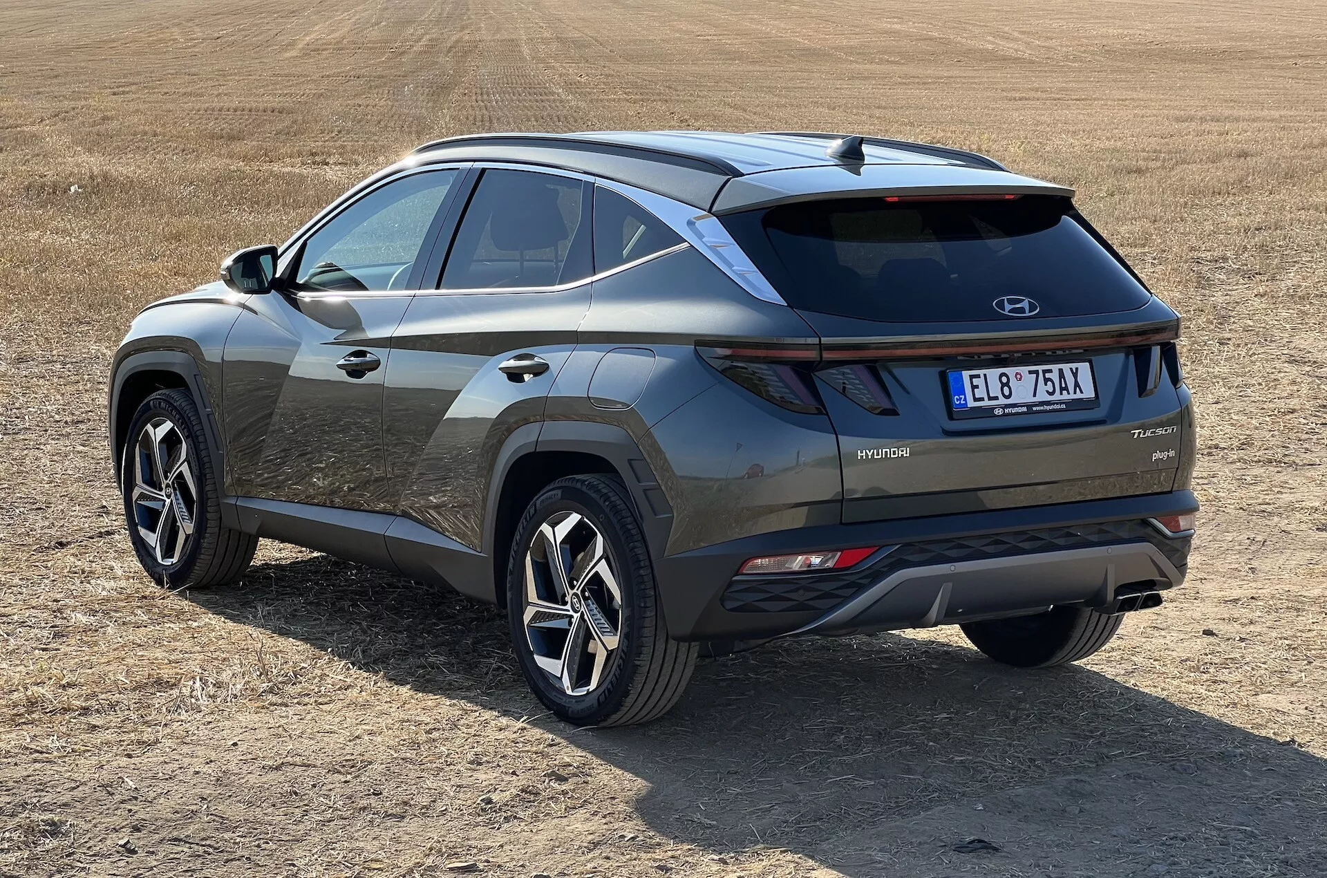 SUV moderne Hyundai Tucson 2020 en vue oblique présentant les profils arrière et latéral avec badge de technologie hybride sur champ herbeux sec, mettant en évidence les feux arrière LED, l'insert de pare-chocs contrastant et les roues sportives à multiples branches