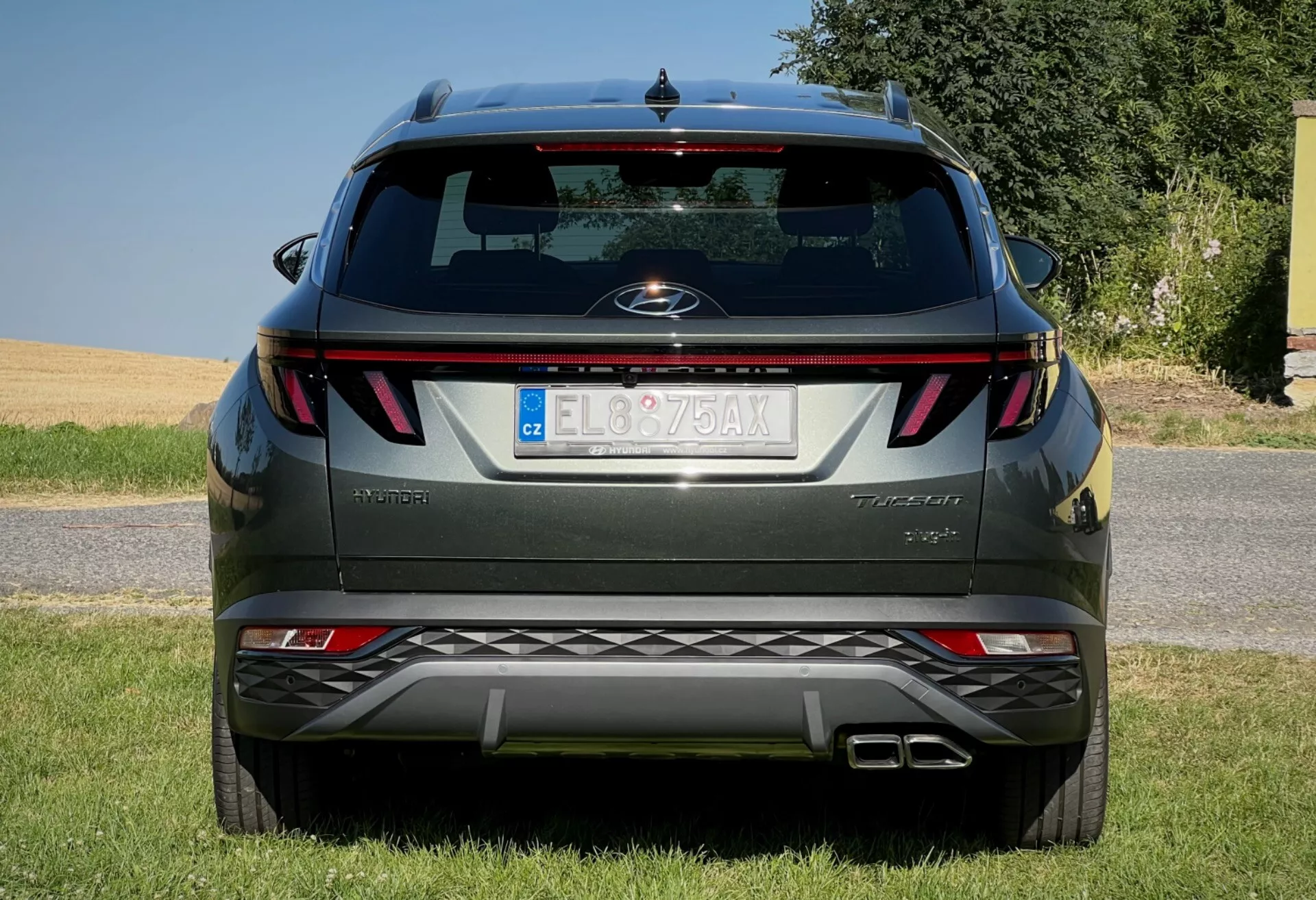 Vue arrière du Hyundai Tucson 2020 montrant des feux arrière horizontaux, un badge central, le nom du niveau de finition, des capteurs de stationnement intégrés dans le pare-chocs et un système d'échappement double, stationné sur une surface bordée d'herbe contre un ciel dégagé