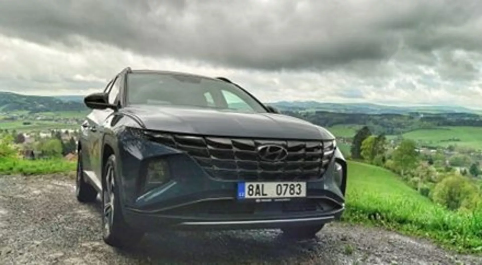 SUV Hyundai Tucson 2020 garé sur un chemin de gravier, montrant sa grille audacieuse, ses phares LED élégants et sa peinture métallique dans une vue dynamique sous un angle avant bas, avec un paysage vallonné luxuriant sous un ciel nuageux en arrière-plan