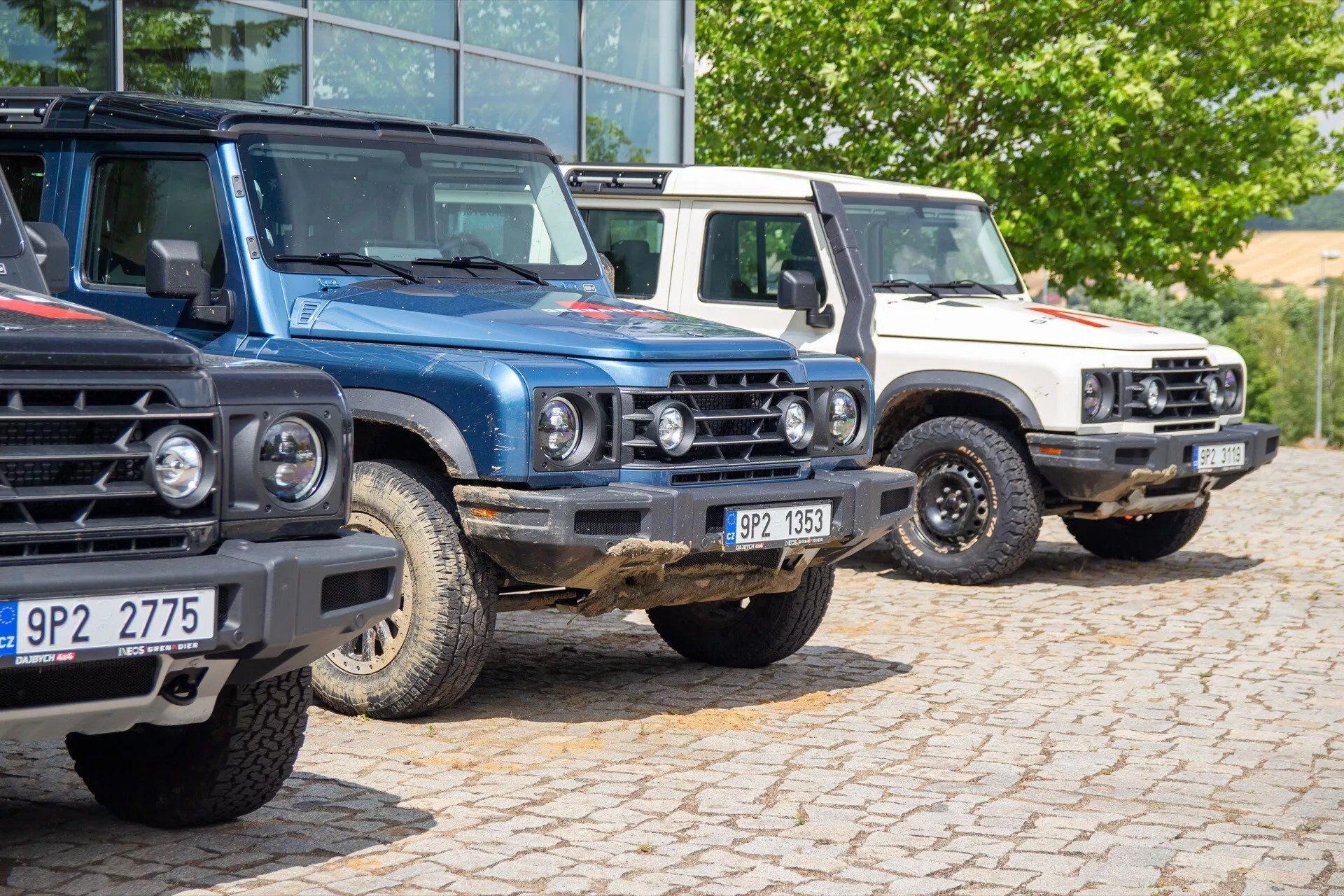Alignement de la première génération d'Ineos Grenadier 2022, comprenant trois SUV avec accessoires tout-terrain stationnés dans un parking, mettant en valeur la grille avant, les phares et un design robuste et carré
