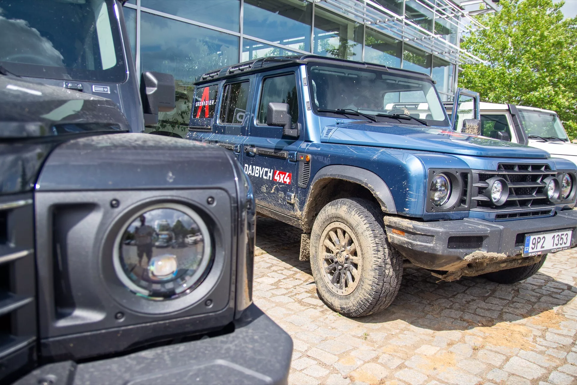 Vue rapprochée de l'Ineos Grenadier 1ère génération 2022 en bleu avec pneus boueux et marquage DAKAR 4X4, démontrant des capacités tout-terrain avec un snorkel, des projecteurs et des réflecteurs de sécurité, vu d'une perspective frontale de trois quarts à côté d'un autre SUV noir