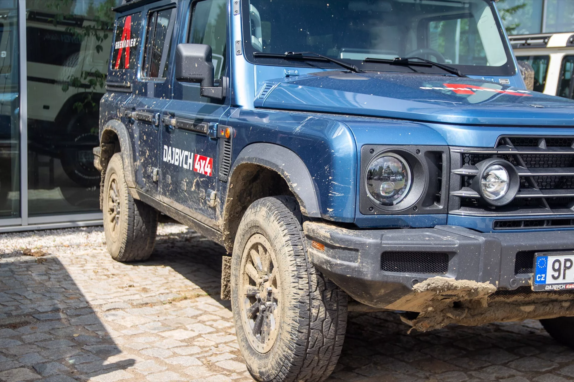 Ineos Grenadier 1ère génération 2022 en bleu poussiéreux présentant perspective frontale latérale avec pneus boueux tout-terrain, snorkel, rétroviseur extérieur robuste, pare-chocs protecteur et marquages du système de transmission intégrale 4x4, soulignant la garde au sol et la posture robuste, prise depuis un angle bas.