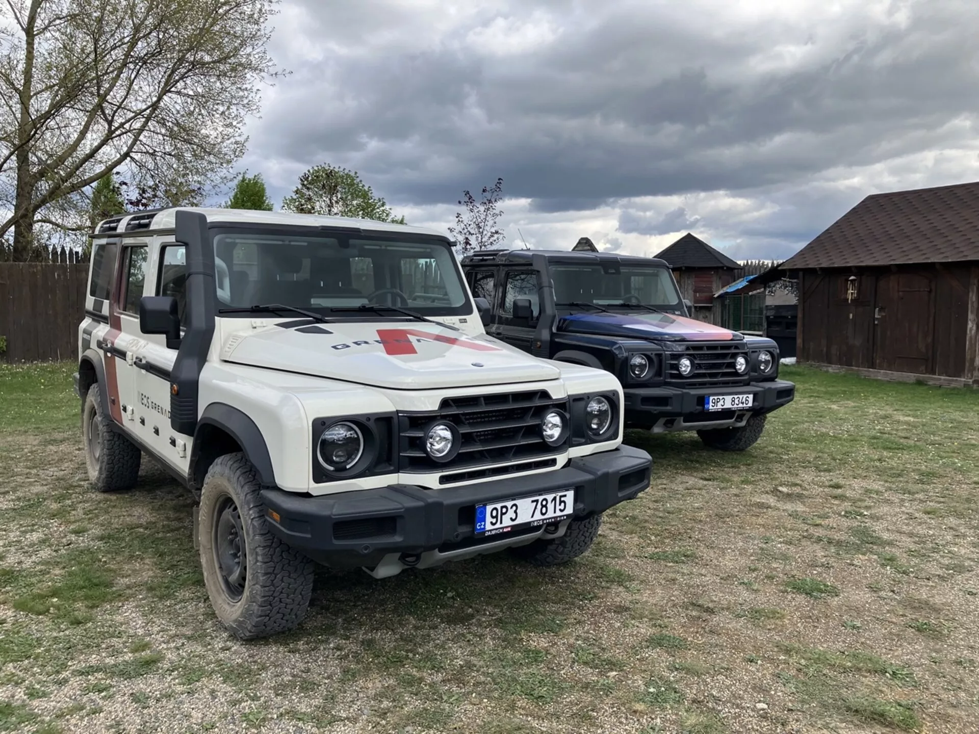 INEOS Grenadier Quartermaster édition 2022 garée en extérieur dans un champ herbeux sous un ciel couvert, présentant un schéma de couleurs bicolores, un snorkel, un porte-bagages avec équipement, une barre lumineuse LED, un emblème de croix rouge d'urgence, un bull-bar avant, des élargisseurs d'aile, et une roue de secours sur le toit, à côté d'un véhicule de conception similaire avec couleur de toit contrastante