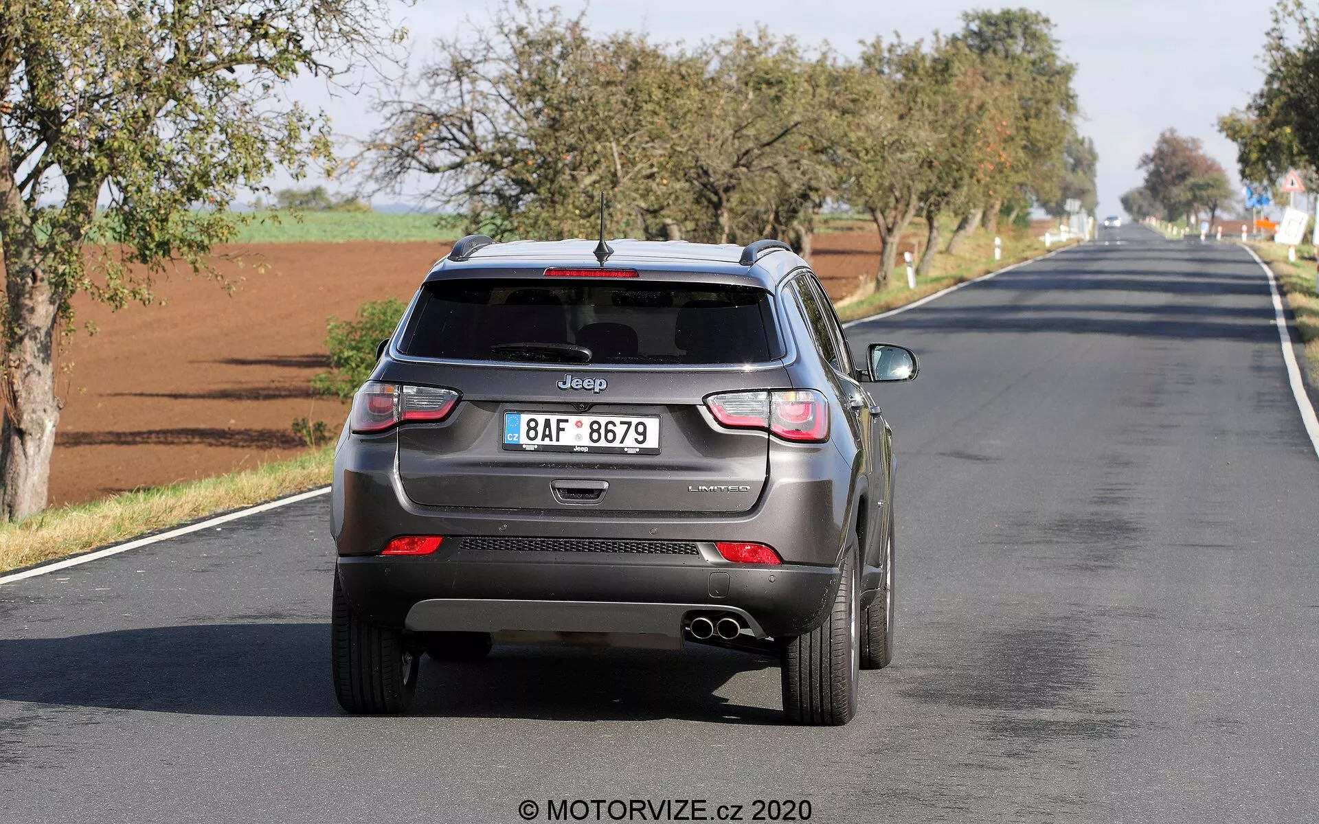 Vue arrière d'une Jeep Compass 2017 s'éloignant sur une route à deux voies, présentant des éléments de design modernes tels qu'une antenne en aileron de requin, un essuie-glace arrière, des feux arrière à LED, des capteurs de stationnement intégrés, des doubles tuyaux d'échappement et une plaque d'immatriculation de style européen avec le logo Jeep clairement visible. Le véhicule semble en mouvement, capturant une utilisation en conditions réelles avec des technologies d'assistance à la conduite potentielles.