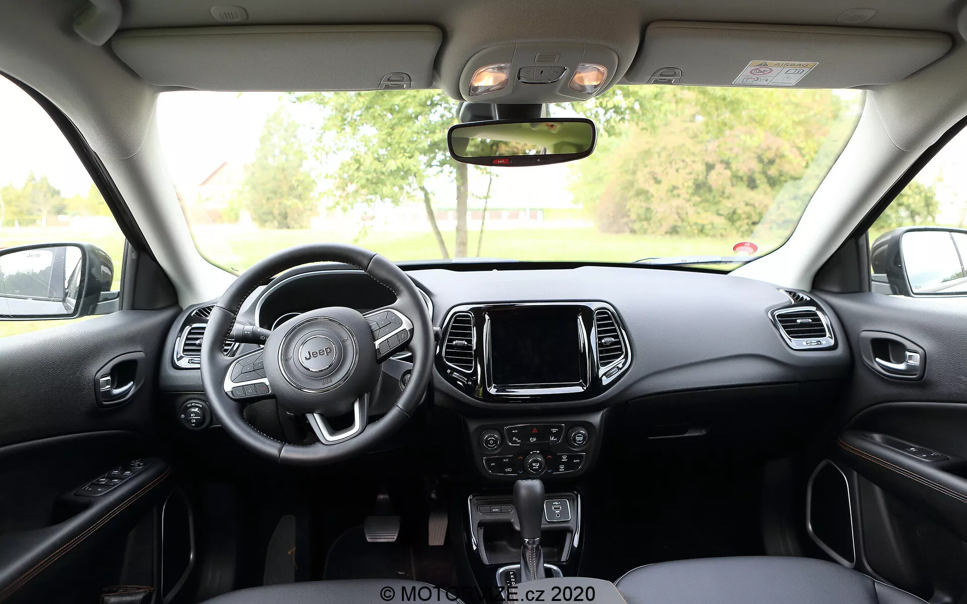 Vue intérieure de la Jeep Compass 2017 depuis la banquette arrière, montrant un volant à trois branches avec des commandes intégrées, une console centrale avec un système d'infodivertissement à écran tactile, des commandes manuelles pour l'audio et le climat en dessous, et un levier de boîte de vitesses automatique. Le tableau de bord et les sièges avant présentent un schéma bicolore avec des accents métalliques ou plastiques, tandis qu'au-dessus se trouvent des commandes de lumière de cabine et de toit ouvrant.