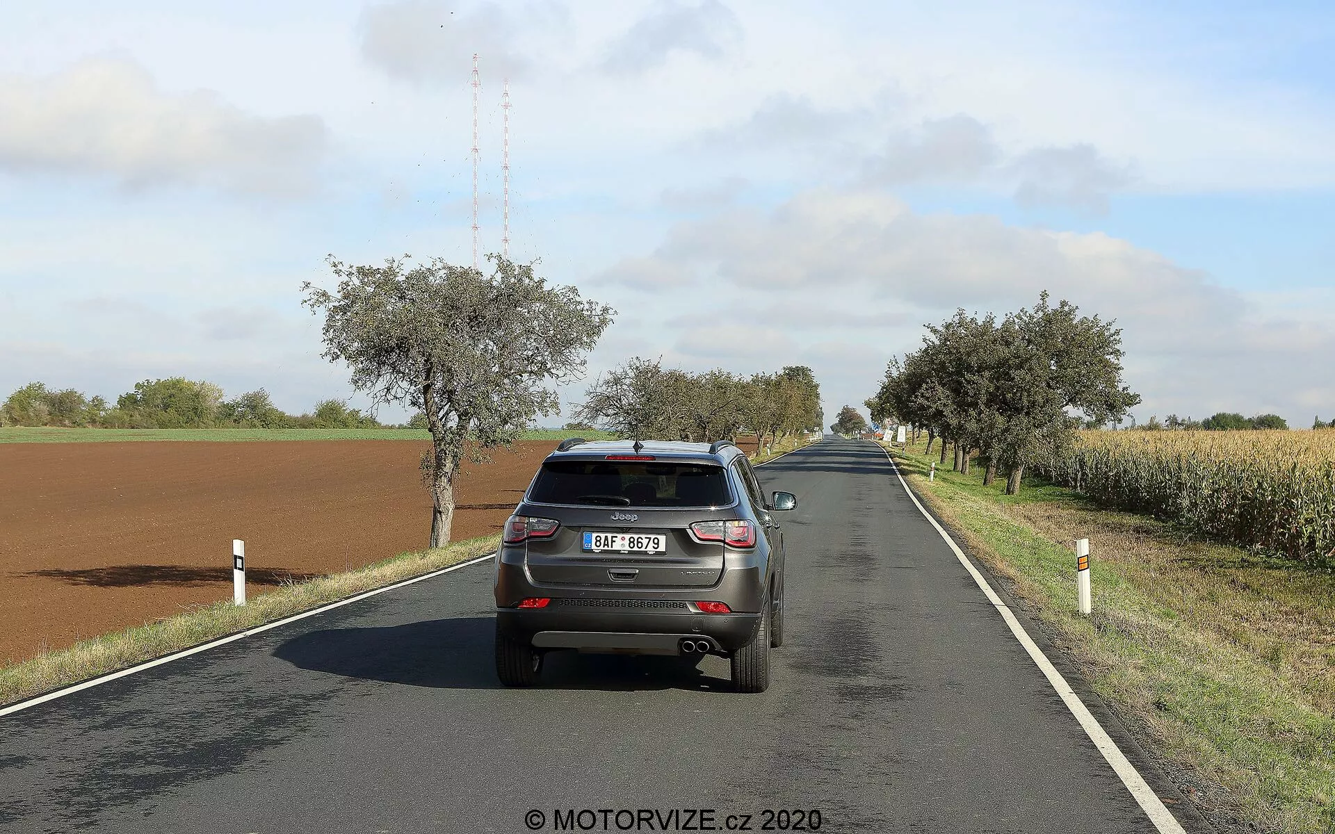 L'image montre la vue arrière d'une Jeep Compass de 2017 roulant sur une route de campagne à deux voies par une journée ensoleillée avec des nuages épars. Le véhicule, positionné sur le côté droit de la voie, affiche son hayon, son pare-chocs, ses feux arrière, sa lunette arrière, sa plaque d'immatriculation, une partie du toit avec l'antenne en forme de nageoire de requin et la partie supérieure des pneus arrière. La voiture occupe une partie significative du cadre, permettant une visualisation claire de ses caractéristiques. La route s'incurve doucement à droite au loin, entourée de champs, d'arbres et de poteaux de route. Les feux de freinage de la voiture ne sont pas allumés, suggérant une croisière normale.