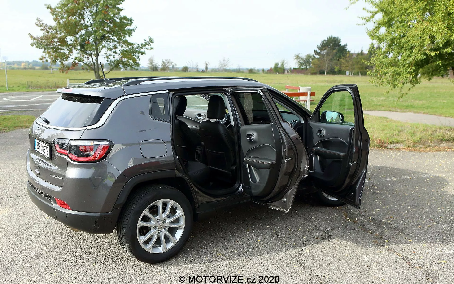 Image d'un SUV Jeep Compass 2017 de taille moyenne garé sur une surface en asphalte dans un cadre ressemblant à un parc avec de l'herbe et des arbres en arrière-plan. La photo est prise sous un angle arrière trois-quarts, montrant le profil et l'arrière du véhicule. Les deux portes côté passager sont ouvertes, révélant l'intérieur en tissu ou cuir sombre, incluant les sièges avant et arrière, avec des caractéristiques potentielles telles que fenêtres et rétroviseurs électriques. Le SUV a une finition métallisée grise et est équipé de roues en alliage à cinq branches, ce qui indique peut-être un niveau de finition supérieur ou une mise à niveau optionnelle des roues. Le filigrane 'MOTORVIZE.cz 2020' suggère que l'image provient d'un site de critique automobile ou d'un site web dédié à l'automobile.