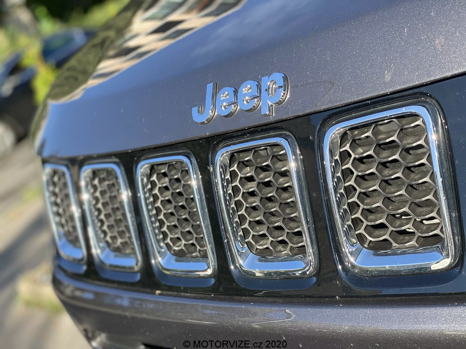 Vue rapprochée de la calandre avant d'une Jeep, montrant le badge 'Jeep' proéminent centré au-dessus des sept fentes verticales, un élément de design signature. La photographie est prise sous un angle oblique, se concentrant de près sur le côté gauche de la calandre, vu d'une perspective légèrement inférieure. Les fentes de la calandre présentent un motif en nid d'abeilles, suggérant une meilleure circulation de l'air et une protection pour le radiateur et les composants du moteur. La finition de peinture noire brillante indique un véhicule relativement neuf ou bien entretenu. La faible profondeur de champ garde le badge 'Jeep' et quelques fentes en focus net, alors que le reste devient flou, soulignant les éléments de design et de marque sous un éclairage naturel, en extérieur, avec des ombres douces.