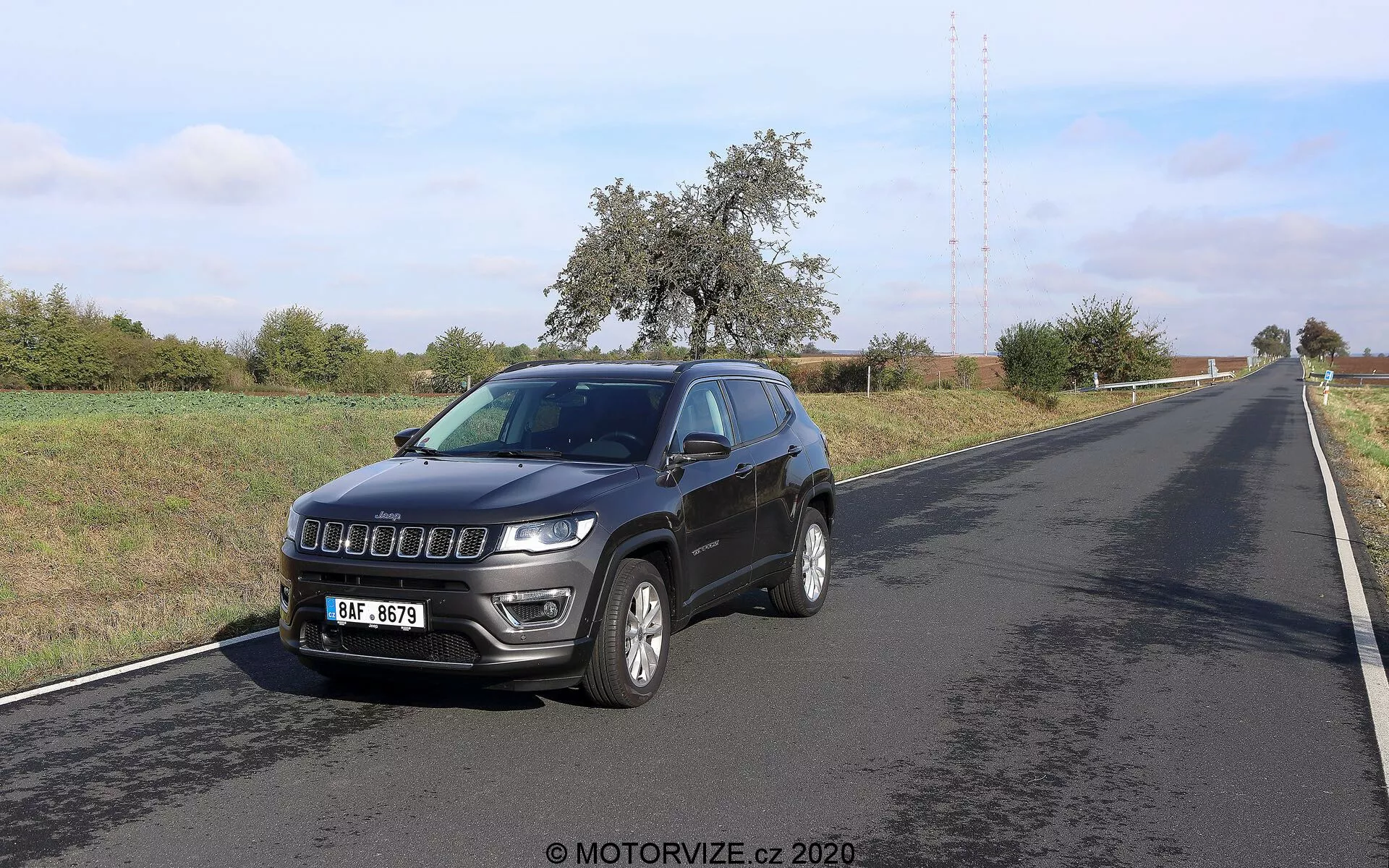 Image d'une Jeep Compass 2017 photographiée sous un angle avant trois quarts. Le véhicule, positionné sur le côté droit d'une route à deux voies, montre son profil avant et latéral. Les caractéristiques clés incluent une calandre moderne, des phares proéminents avec feux de jour à LED, des roues en alliage, des indicateurs de direction intégrés aux rétroviseurs latéraux, une antenne de toit et des phares antibrouillard sur le pare-chocs avant. L'environnement semble rural avec un ciel dégagé et de la végétation le long de la route.