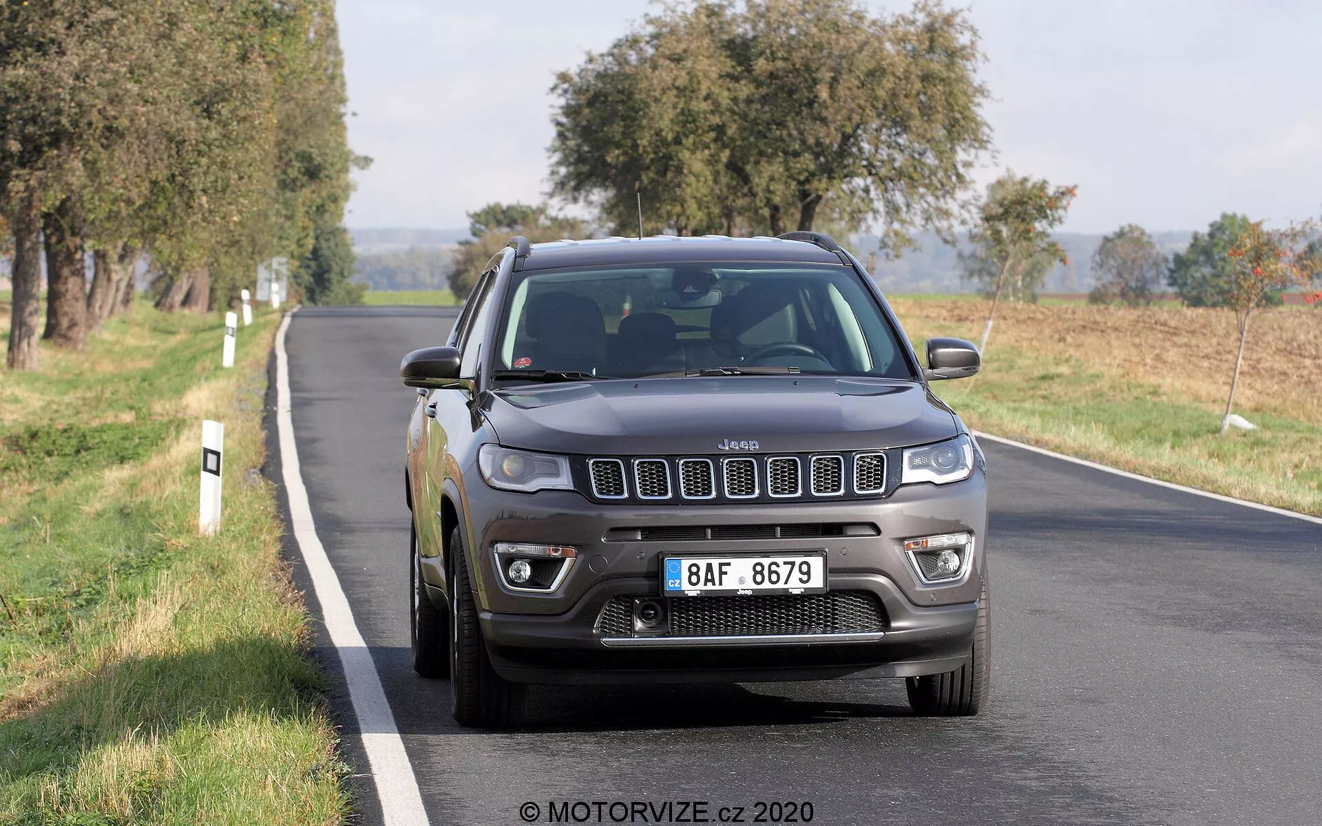 Image d'une Jeep Compass 2017 roulant sur une route à deux voies, photographiée sous un angle de trois quarts avant. La grille à sept fentes emblématique du véhicule, les phares détaillés avec feux de jour à LED, les antibrouillards et les roues en alliage multi-branches sont clairement visibles. La couleur gris foncé ou noire de la Jeep lui donne une apparence solide et robuste, avec la lumière naturelle du jour mettant en évidence ses caractéristiques. Le véhicule semble être en mouvement, avec un léger flou sur les roues. Les arbres et les champs ouverts en arrière-plan suggèrent un cadre rural ou semi-rural. La voiture occupe la moitié centrale et inférieure du cadre, légèrement à droite, capturant sa grille avant, ses phares, son pare-chocs, son capot, son aile côté conducteur, sa roue et sa porte avant.