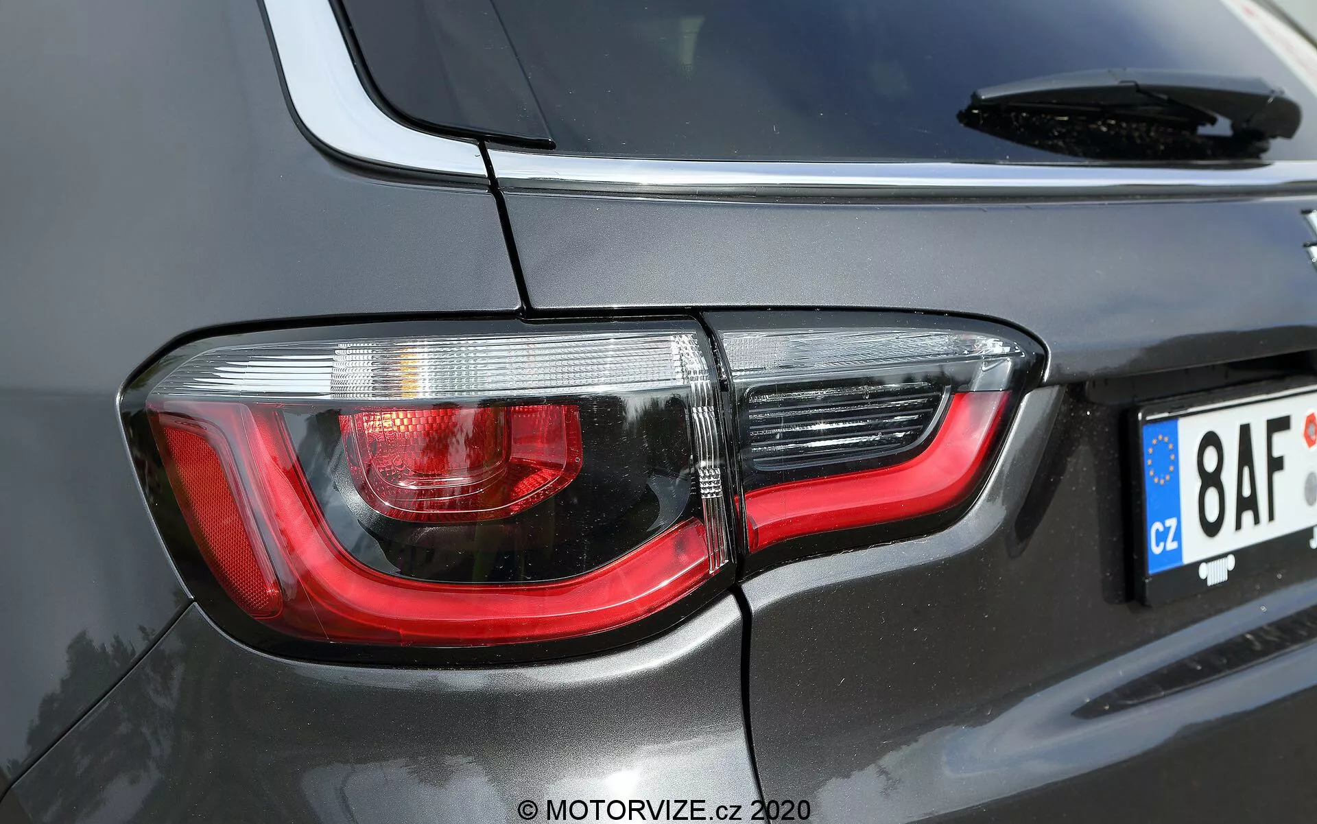 Gros plan sur la section arrière droite d'une Jeep Compass 2017 noire ou gris foncé, mettant en évidence un design moderne de feux arrière avec des lentilles claires et rouges, une technologie LED et un matériau réfléchissant en chrome pour une meilleure visibilité. L'image montre une partie du pare-brise arrière, l'antenne et une plaque d'immatriculation de style européen, prise sous un angle diagonale et décentré qui souligne la courbure et le style contemporain du SUV.