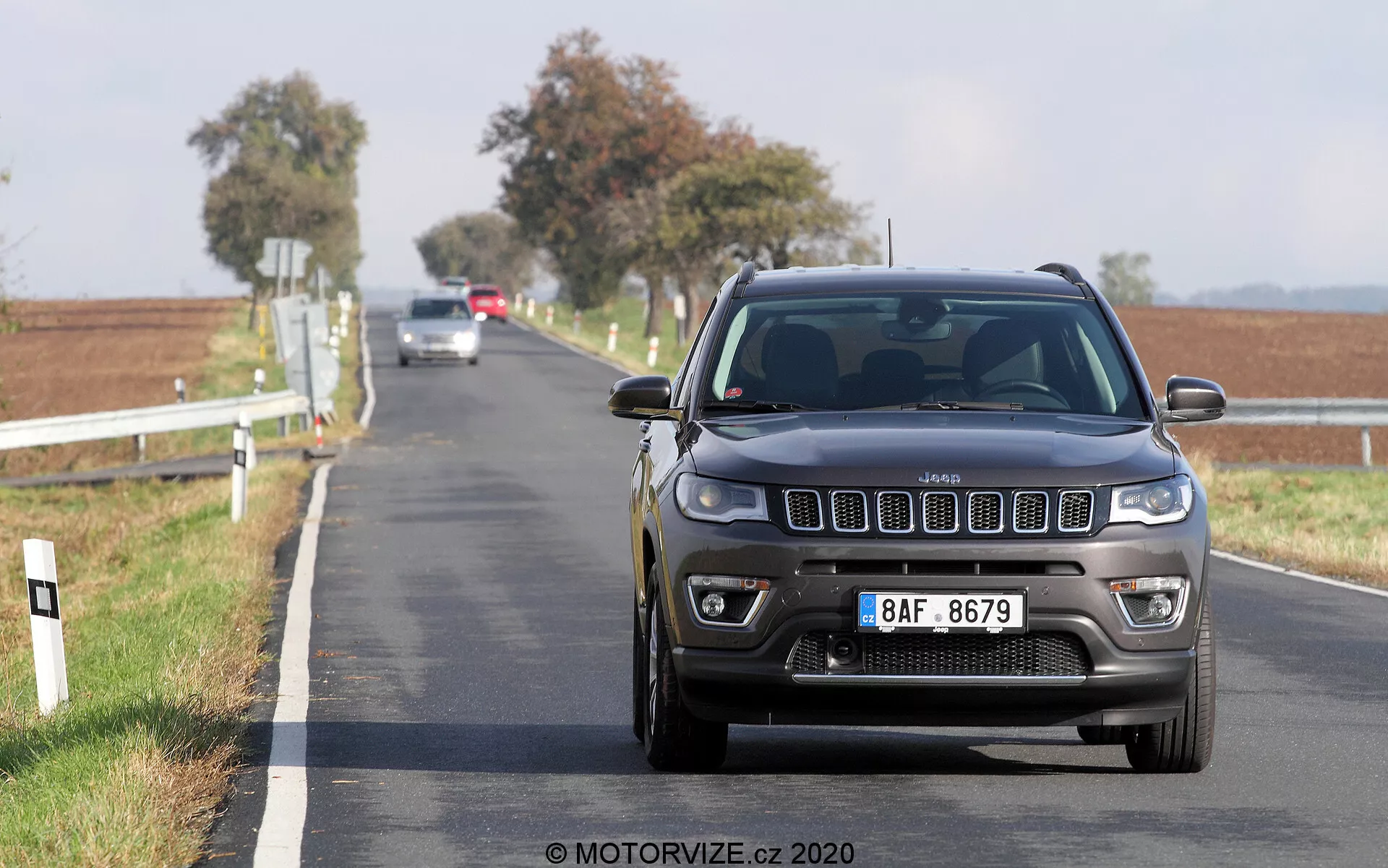Prise de vue en contre-plongée d'une Jeep Compass 2017 roulant vers l'appareil photo sur une route pavée à deux voies. La vue de face montre en détail la calandre, les phares et le pare-chocs avec des caractéristiques modernes visibles comme des feux LED ou HID et des capteurs de stationnement avant. La voiture est en mouvement, avec un arrière-plan rural flou présentant des poteaux blancs en bordure de route et des champs ouverts sous un ciel clair.